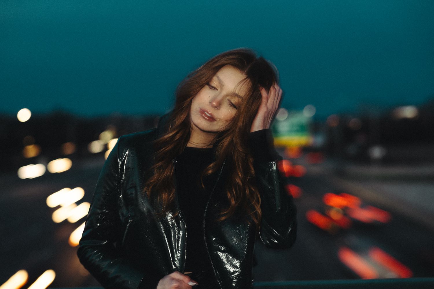 Young woman with long brown hair in leather jacket posing outdoors at night with city lights.