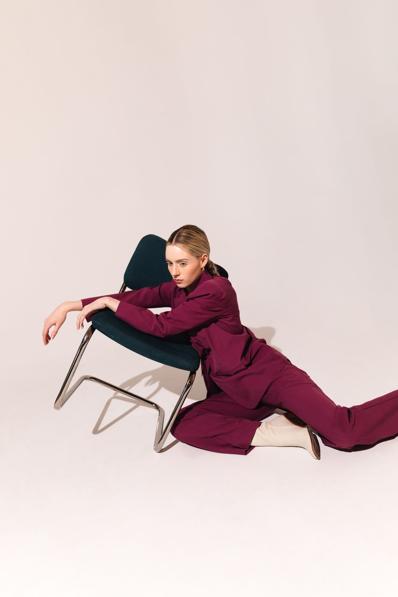 Blonde woman in a burgundy suit reclines against a teal chair on a light gray studio background.