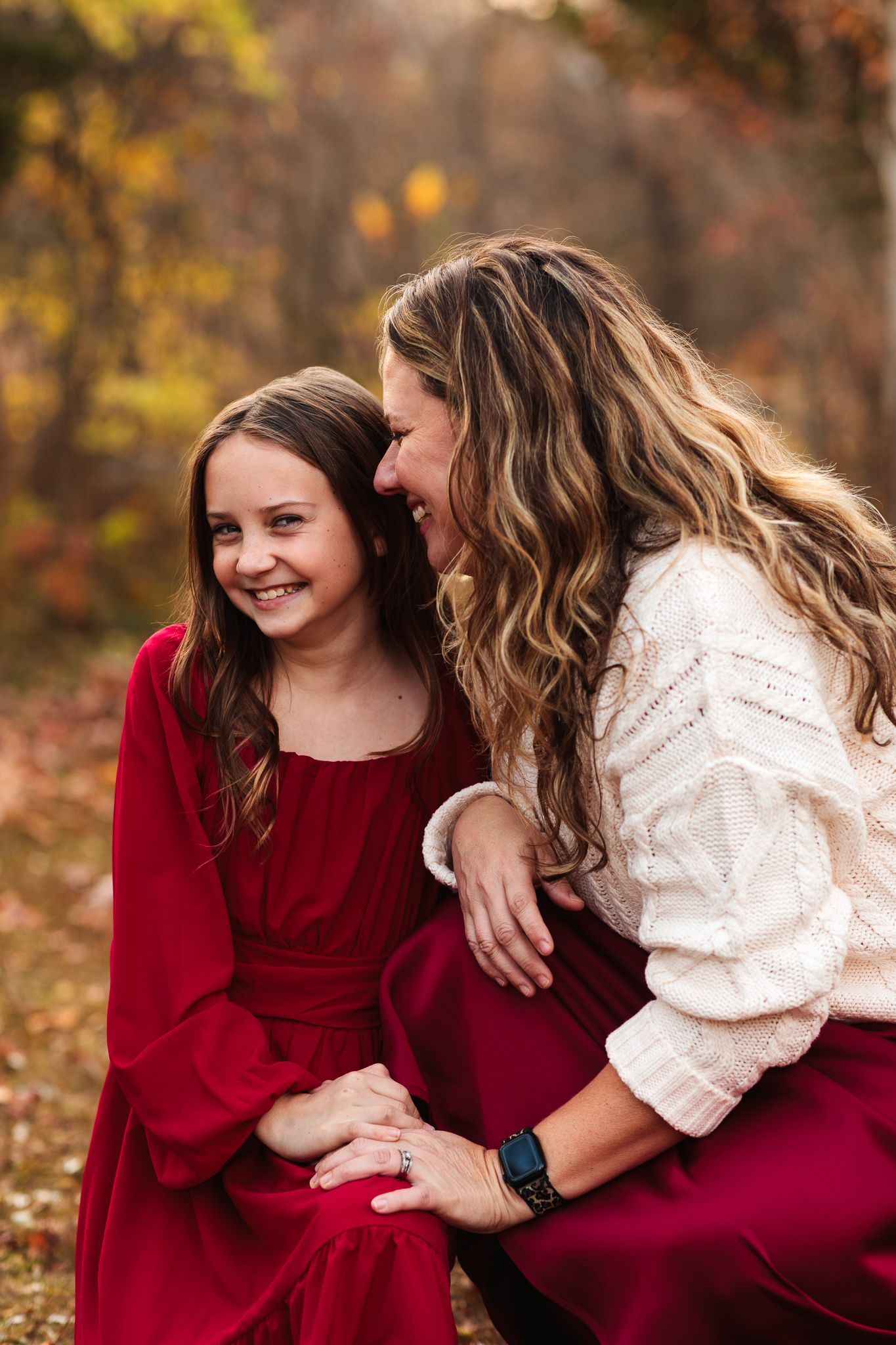 A mother and daughter share a joyful moment outdoors in autumn, both wearing red dresses, surrounded by fall foliage.