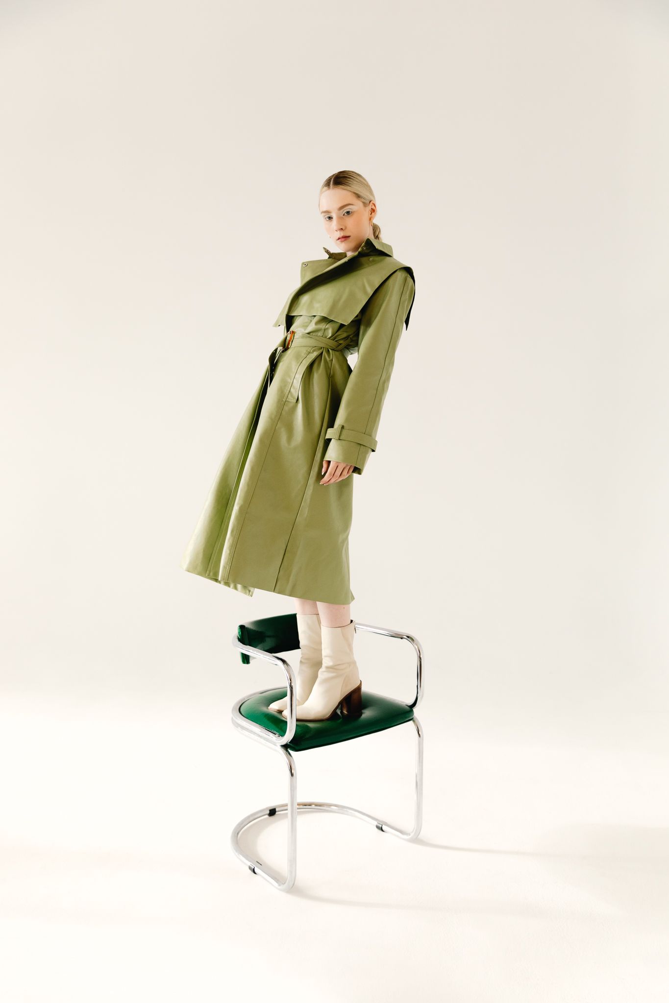 Blonde woman in green trench coat standing on chair wearing cream boots in white studio.