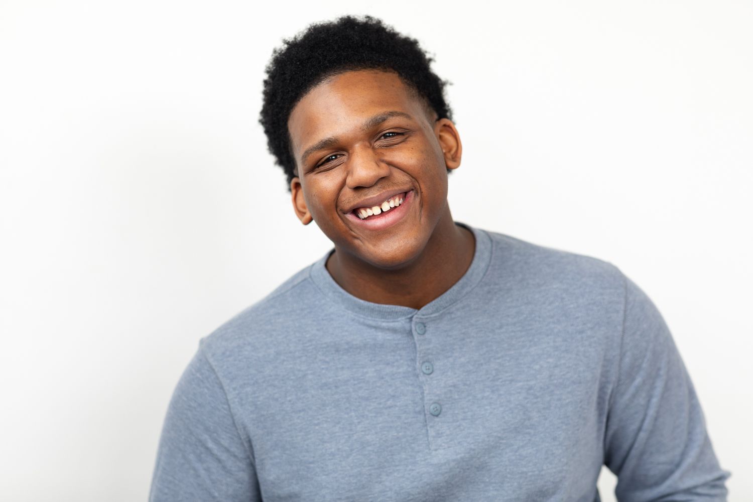 Young Black man laughing in gray henley shirt against white background.