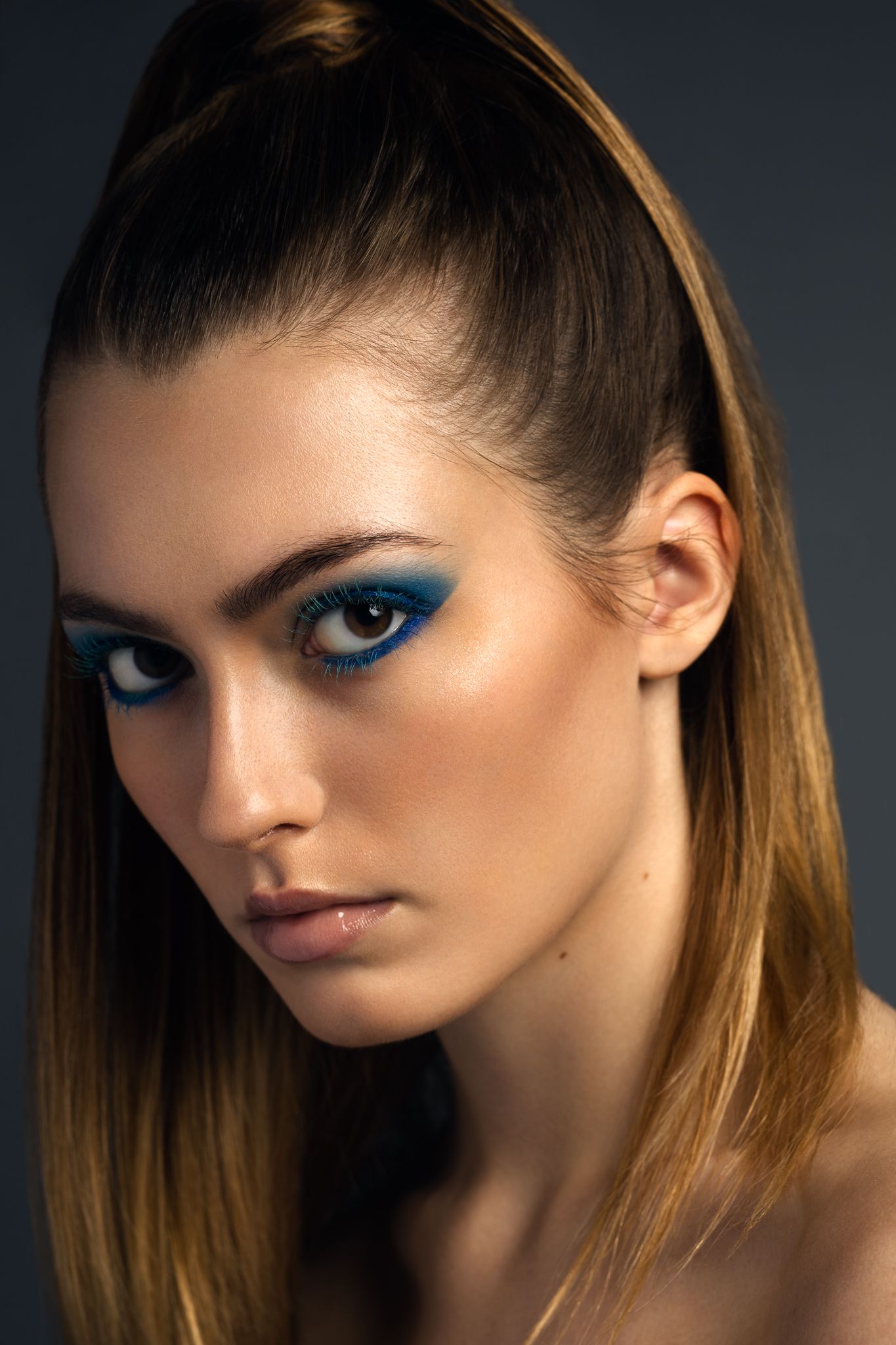 Woman with dramatic blue eye makeup and sleek ponytail posing against dark gray background.