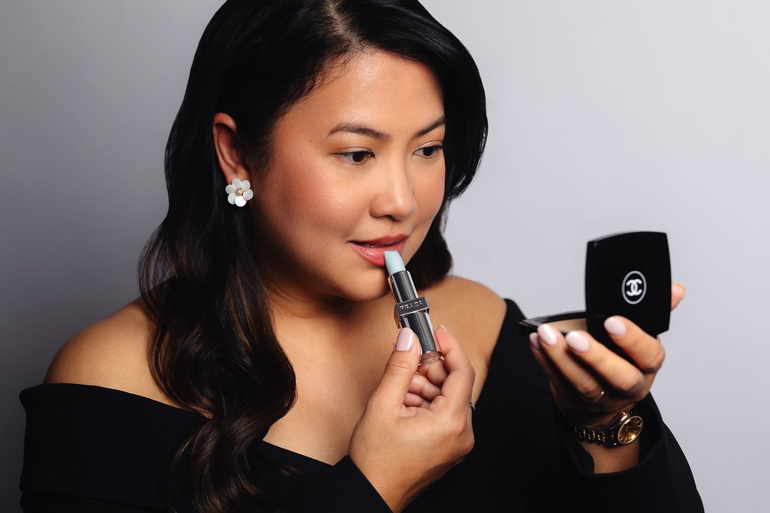 Asian woman applying red Chanel lipstick using a compact mirror, wearing a black off-shoulder top and pearl earrings.