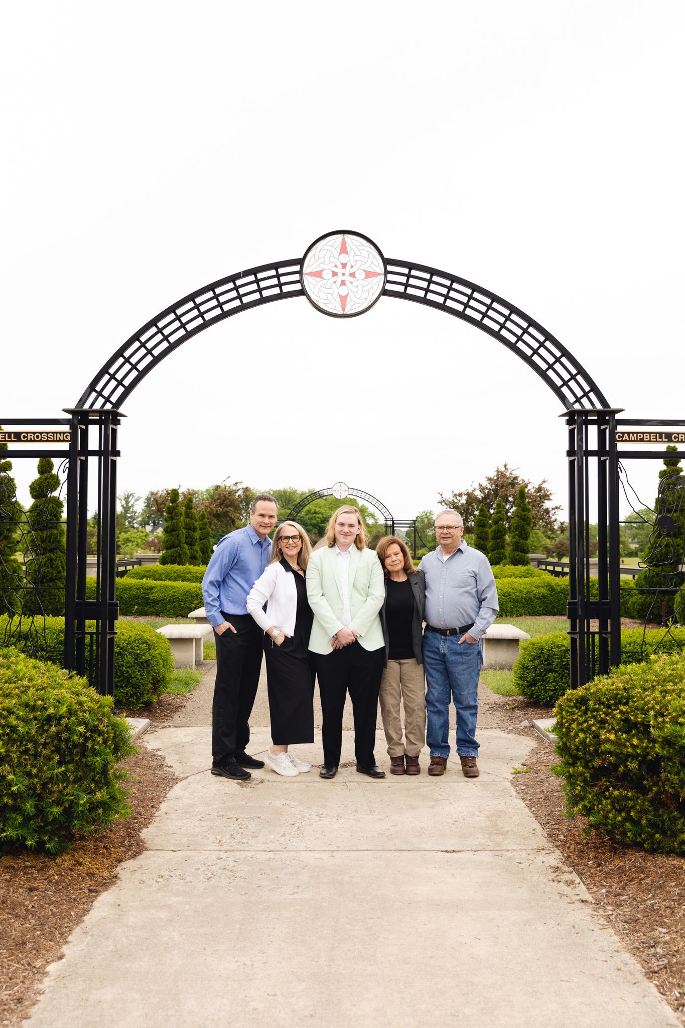 Family of five posing under decorative iron arch in manicured garden park setting.