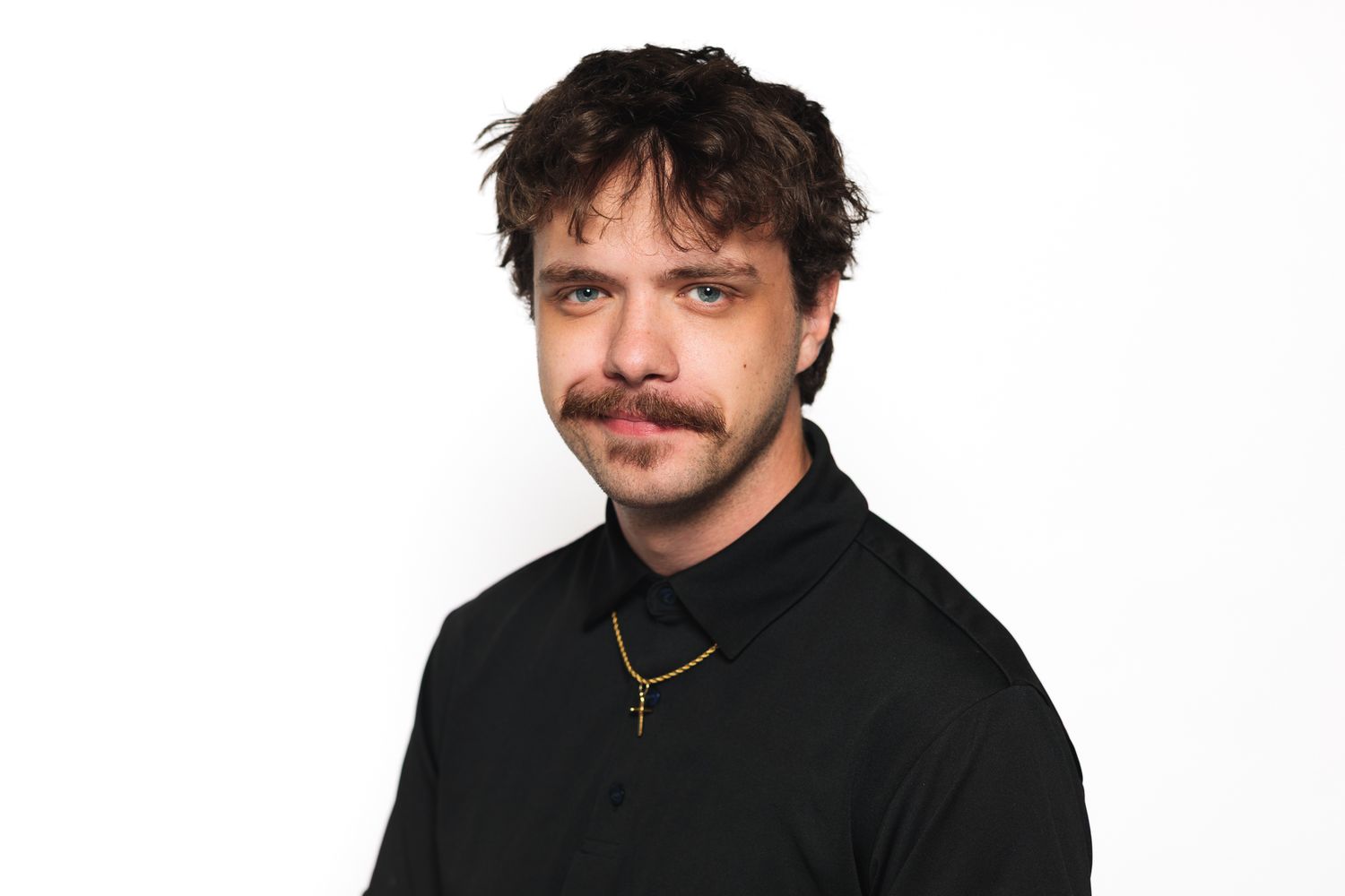 Young man with mustache wearing black shirt and gold cross necklace against white background.