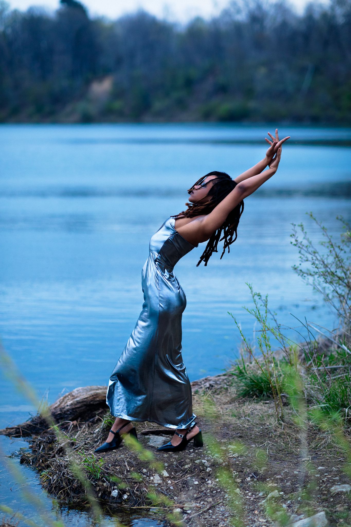 Woman in silver dress dancing gracefully by a blue lake, arms outstretched, with lush greenery in the background.