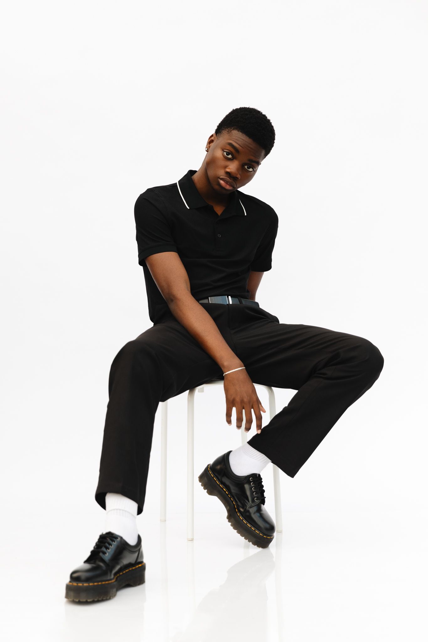 Young Black man in black polo shirt, trousers, and Dr. Martens boots sitting on white stool against white background.