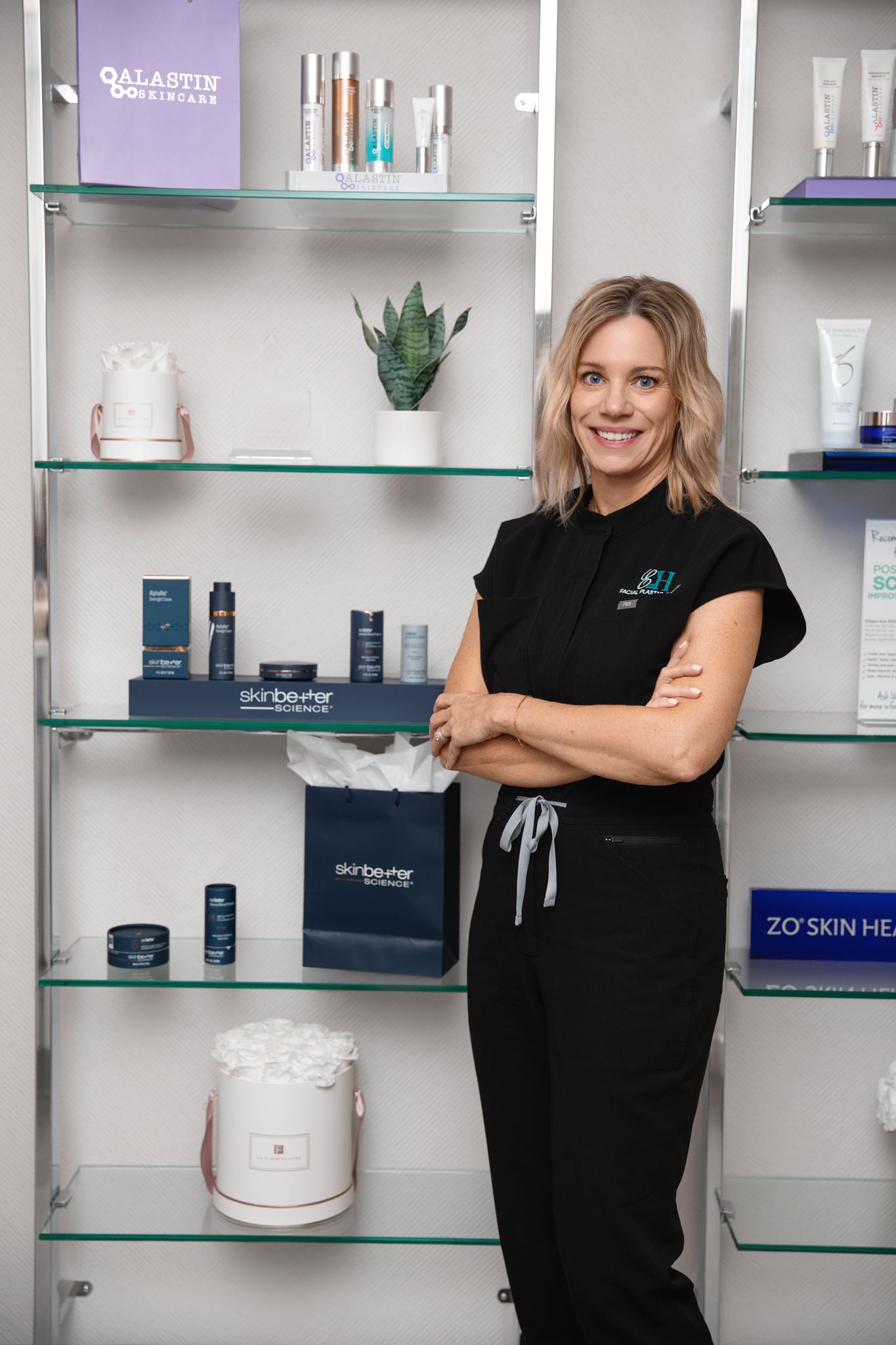 Smiling esthetician in black uniform stands confidently before skincare product shelves featuring Alastin and ZO Skin Health brands.