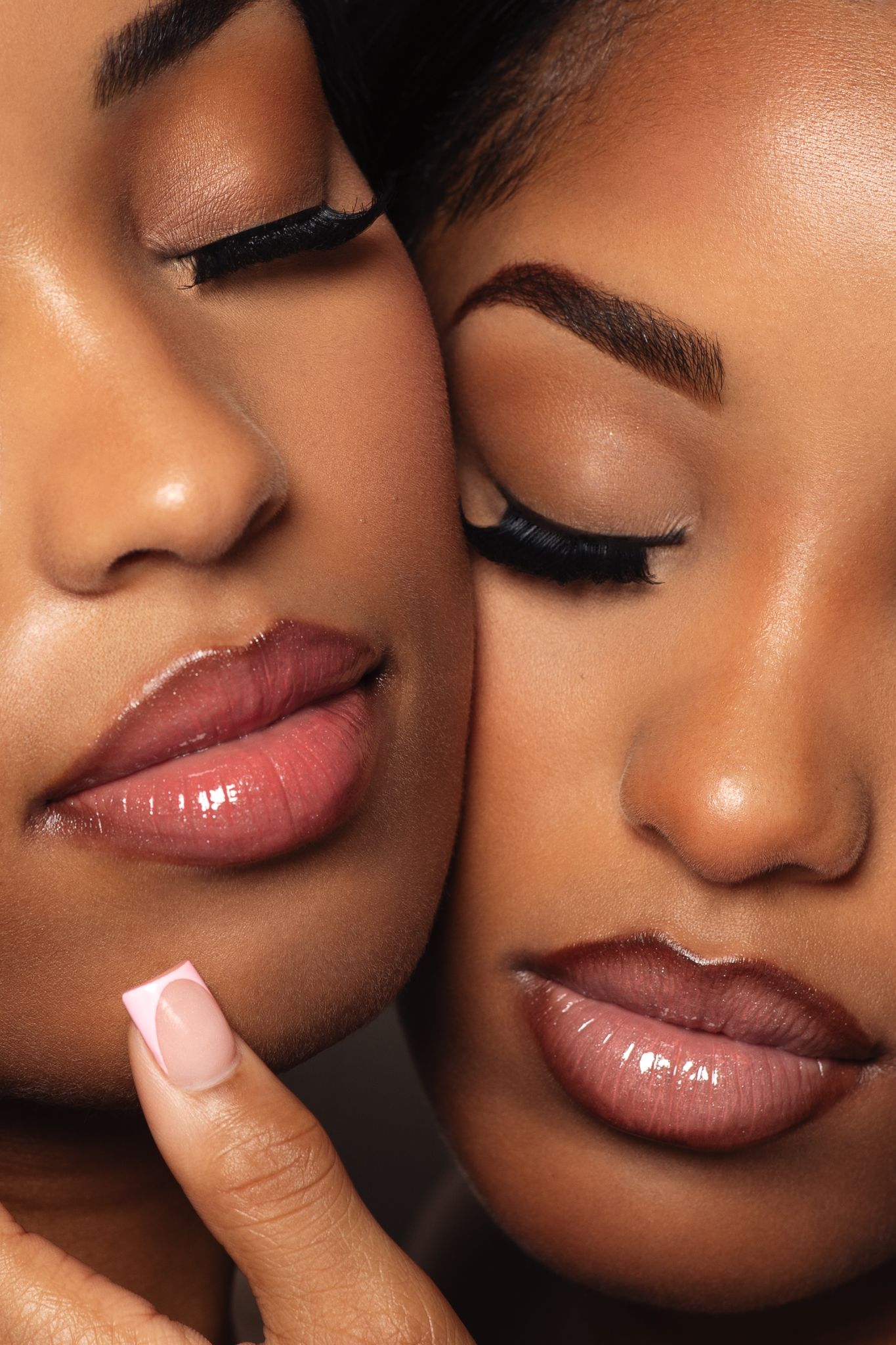 Close-up beauty portrait of two Black women with flawless skin, bold lips, and long lashes facing each other.