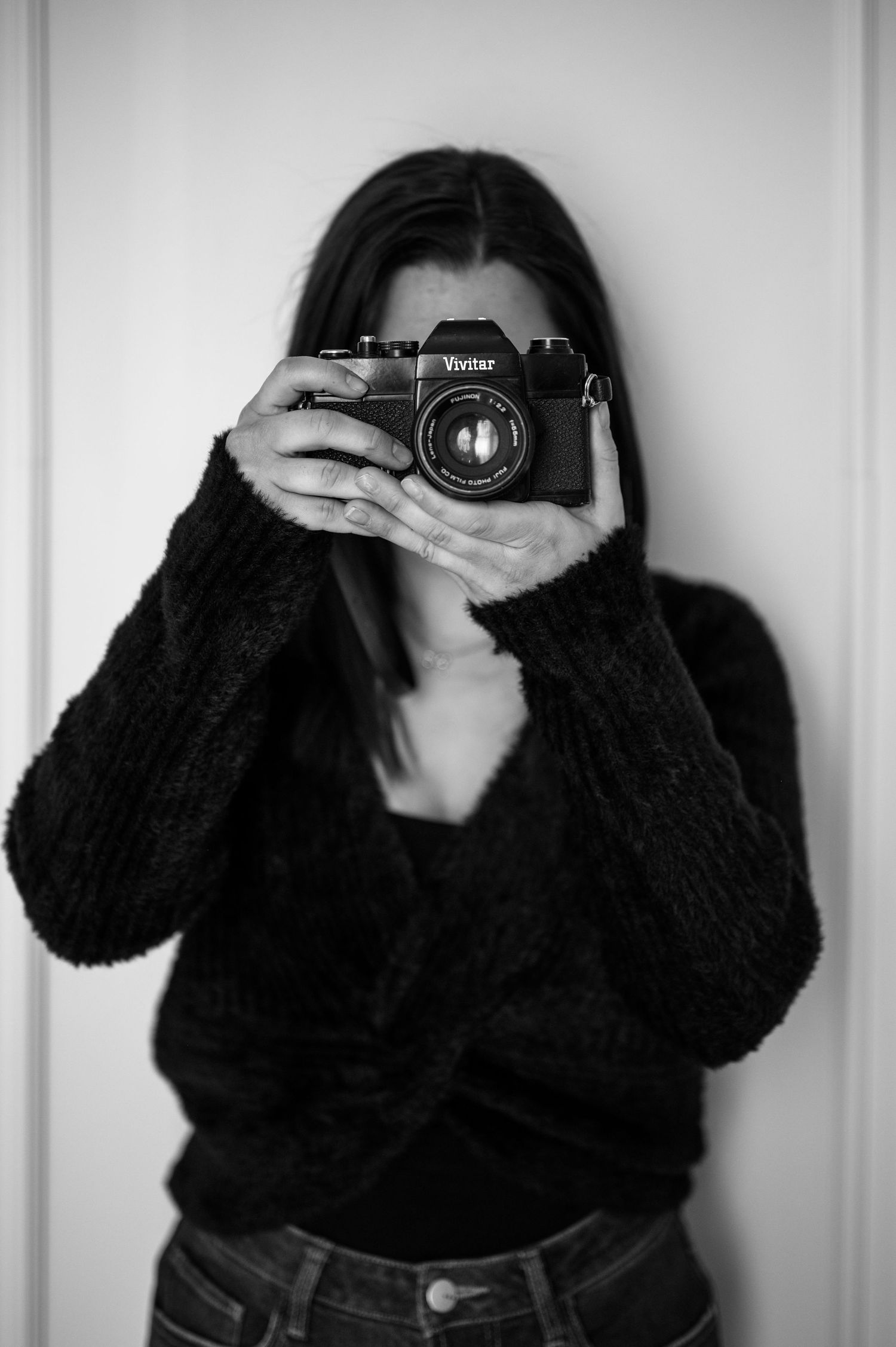 Black and white portrait of a woman holding a Vivitar film camera up to her face in a dark knit sweater.