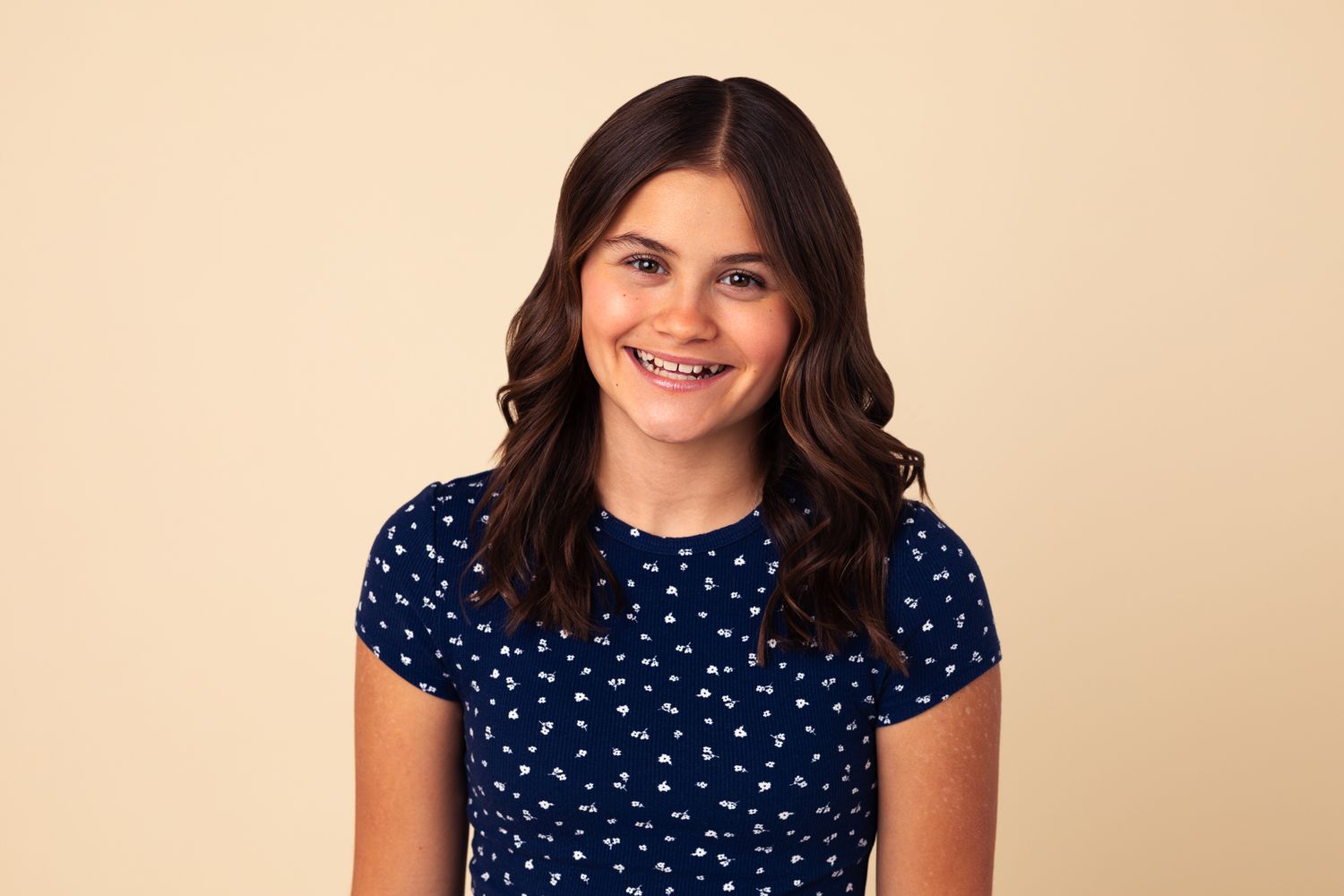 Smiling young girl with braces and dark wavy hair wearing a navy blue polka dot dress against a beige background.
