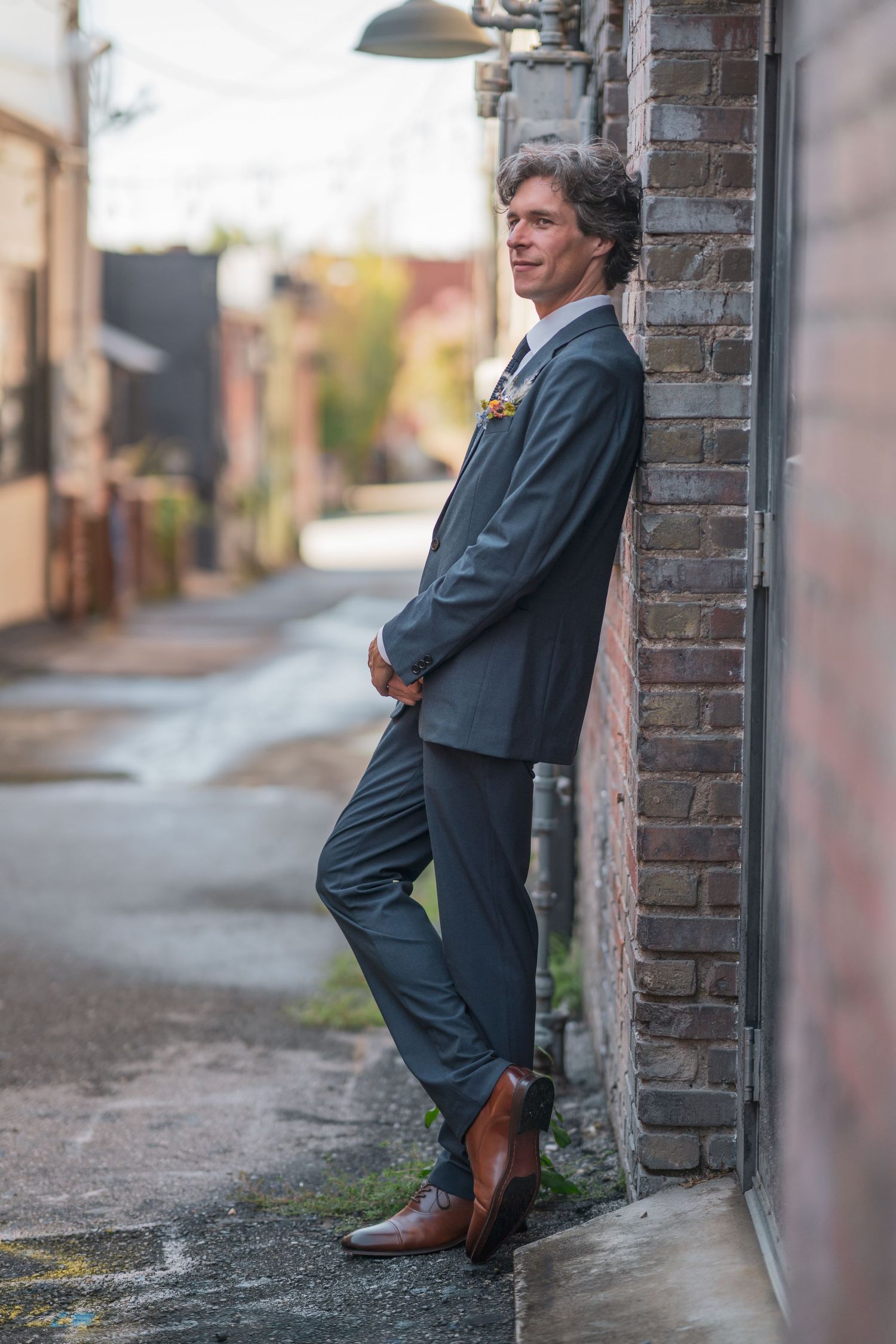A person in a dark suit leans casually against a brick wall in an urban alley setting.