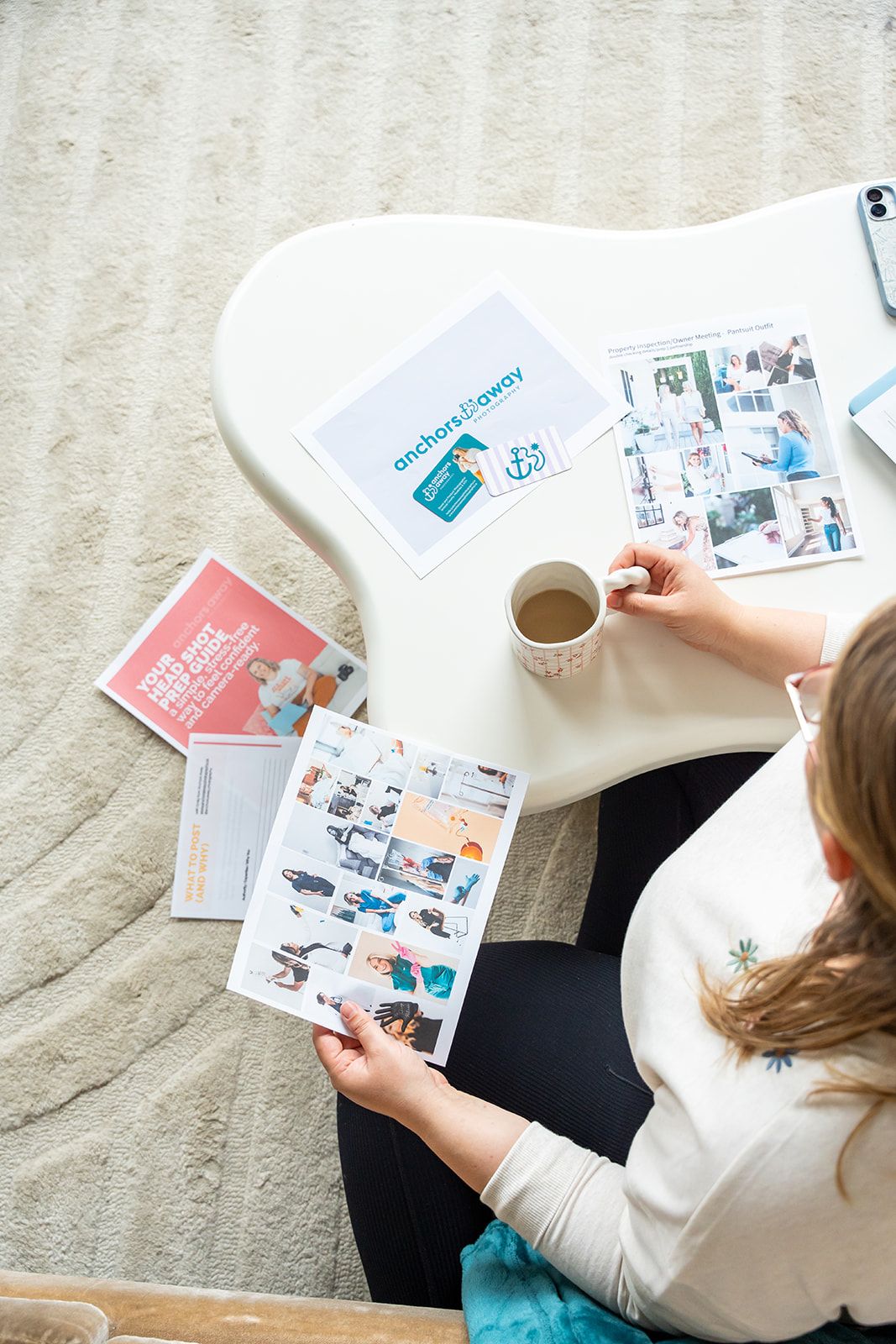 Woman sitting on floor reviewing colorful photo collages and Anchors Away branding materials on a white table.