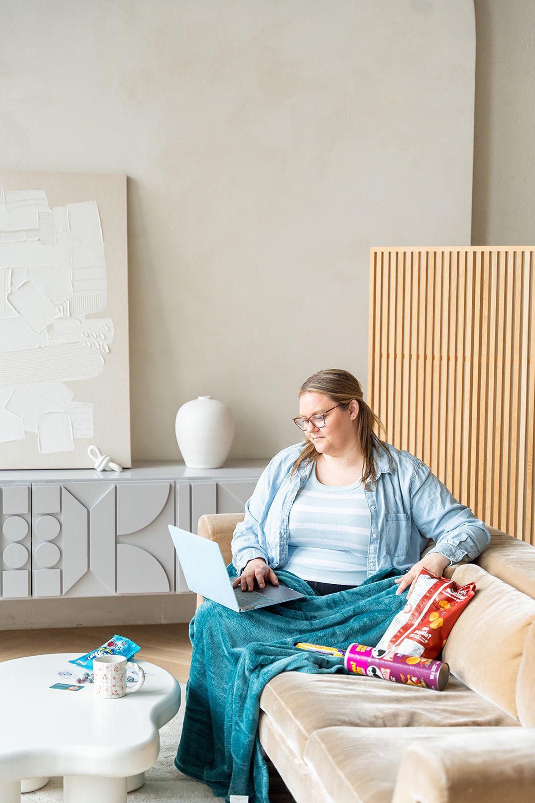 Woman in glasses sits on sofa using laptop while wrapping Christmas gifts in a modern living room.