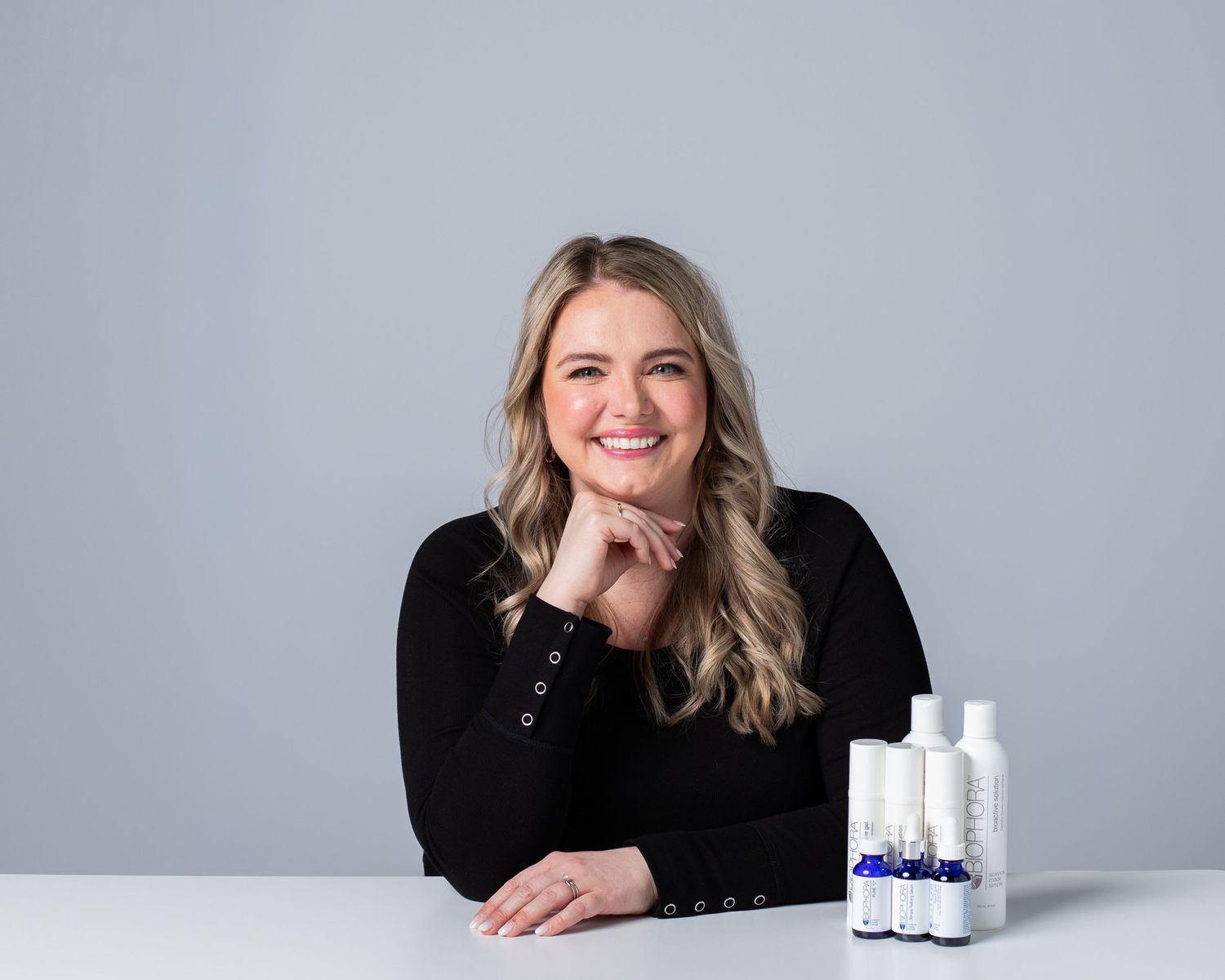 The image shows a smiling woman with long blonde hair sitting at a table with various skincare products.
