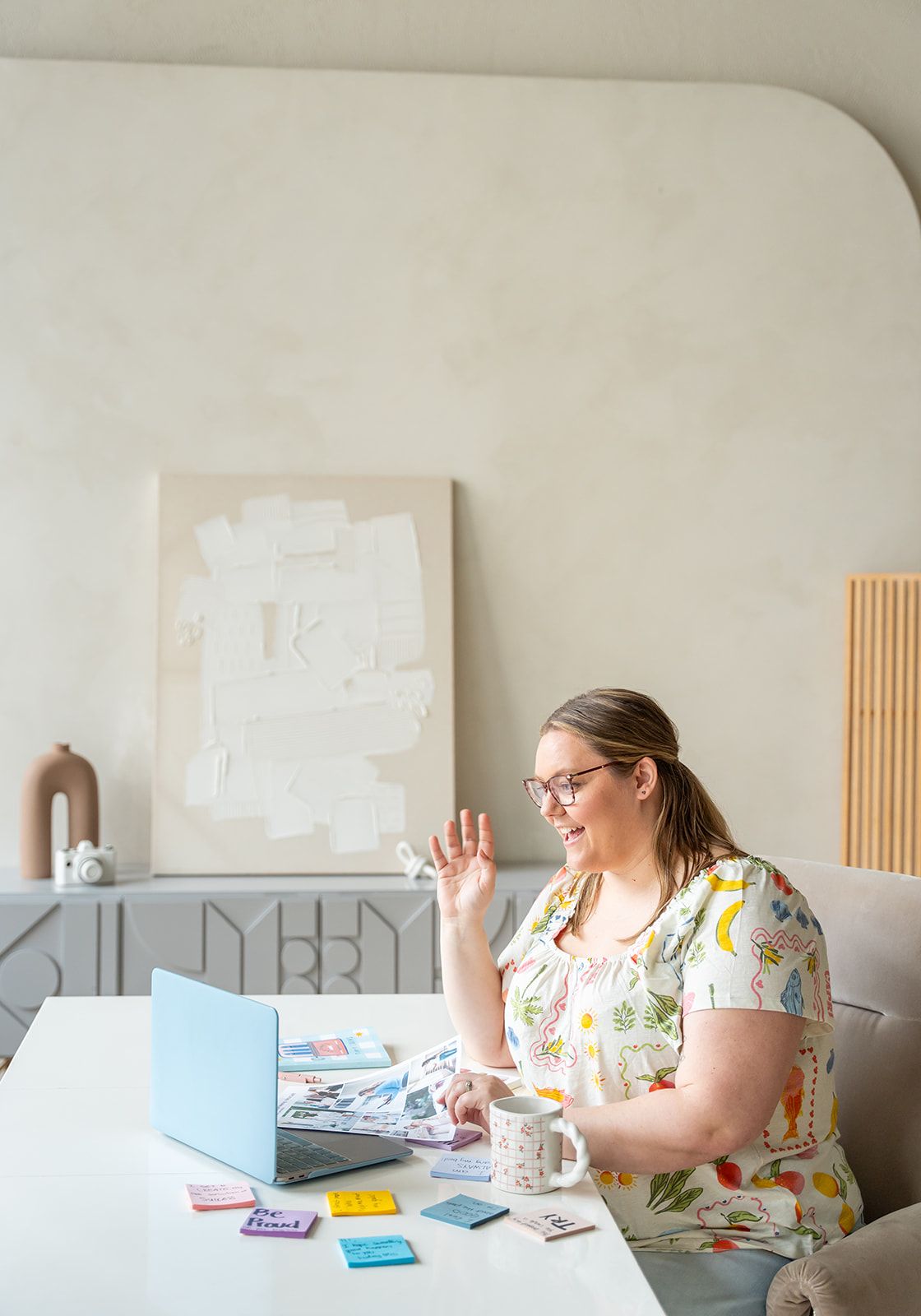 Woman in floral dress waving at laptop during video call, colorful sticky notes on white desk in modern home office.