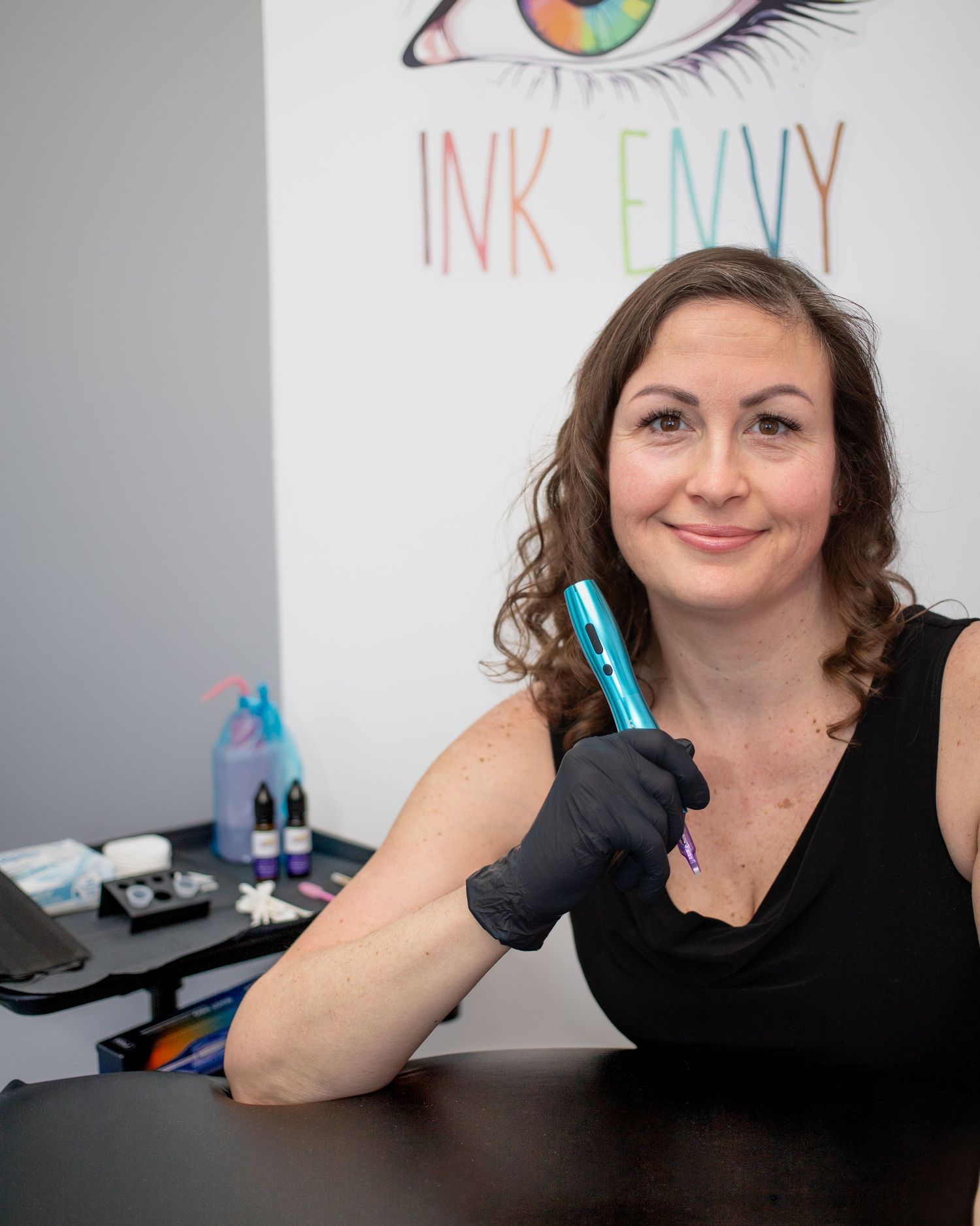 A woman with curly brown hair is holding a tattoo machine and smiling in front of a wall with an eye design.