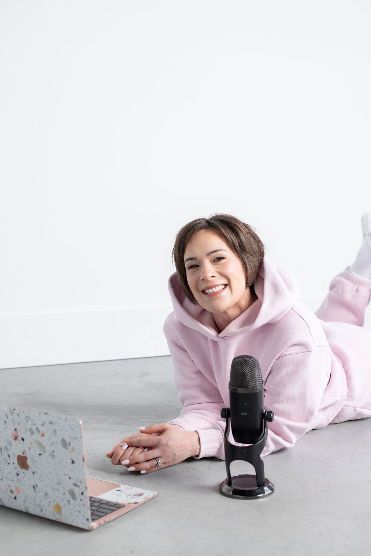 A smiling woman wearing a pink hoodie sits on a couch and holds a microphone.
