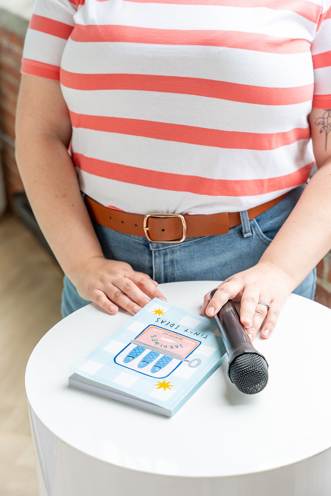 Person in a coral-striped shirt and denim stands at a white round table with a handheld microphone and a light blue notebook.