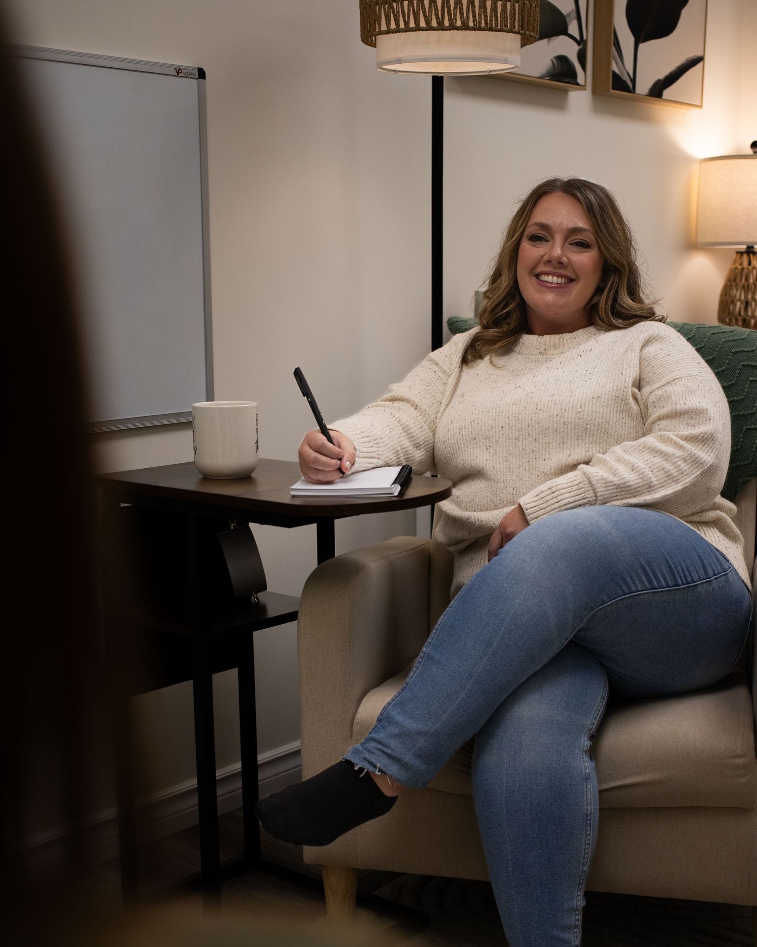 A portrait of a smiling young woman sitting on a chair and writing in a notebook.