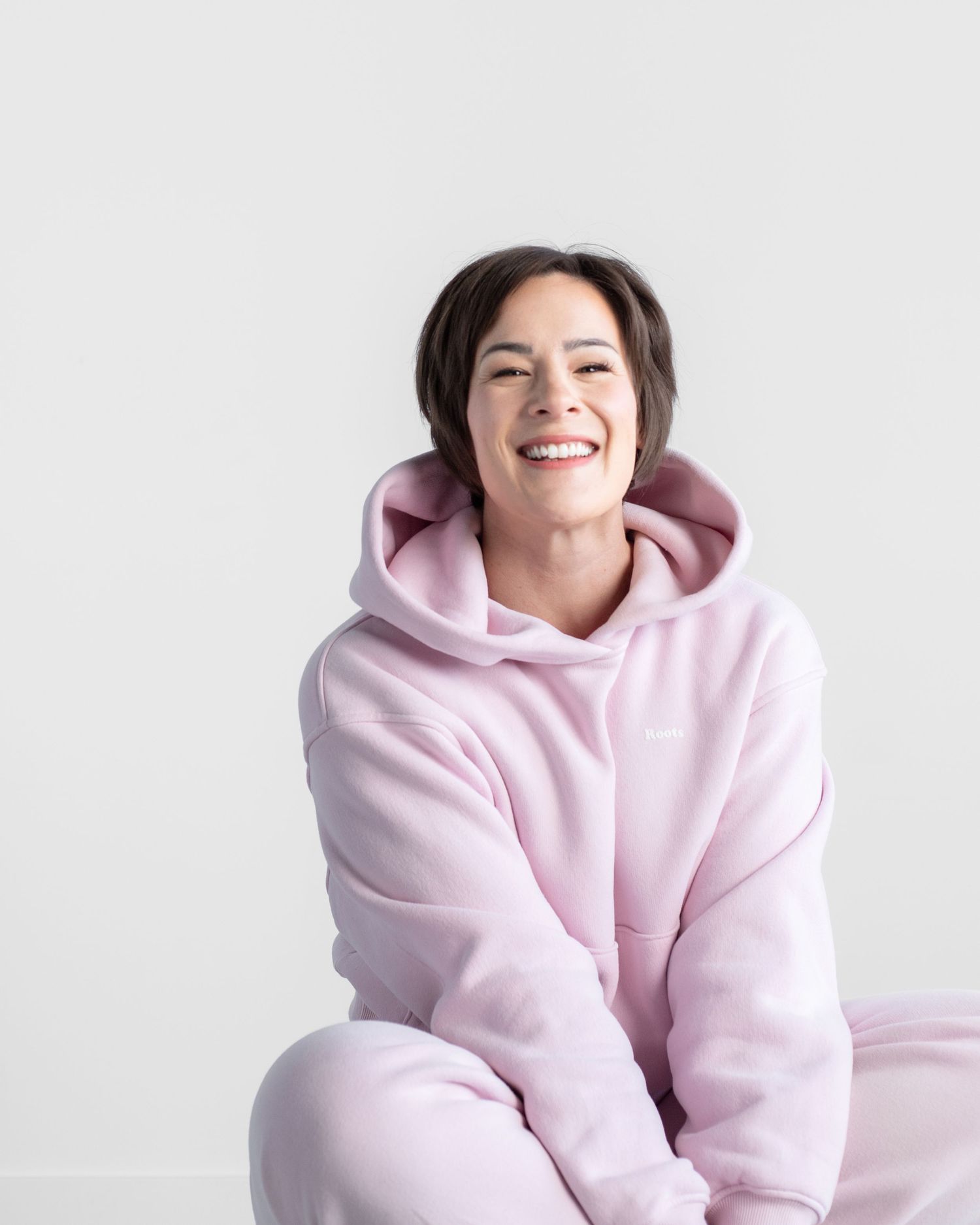 The image shows a smiling woman wearing a pink hooded sweatshirt against a white background.