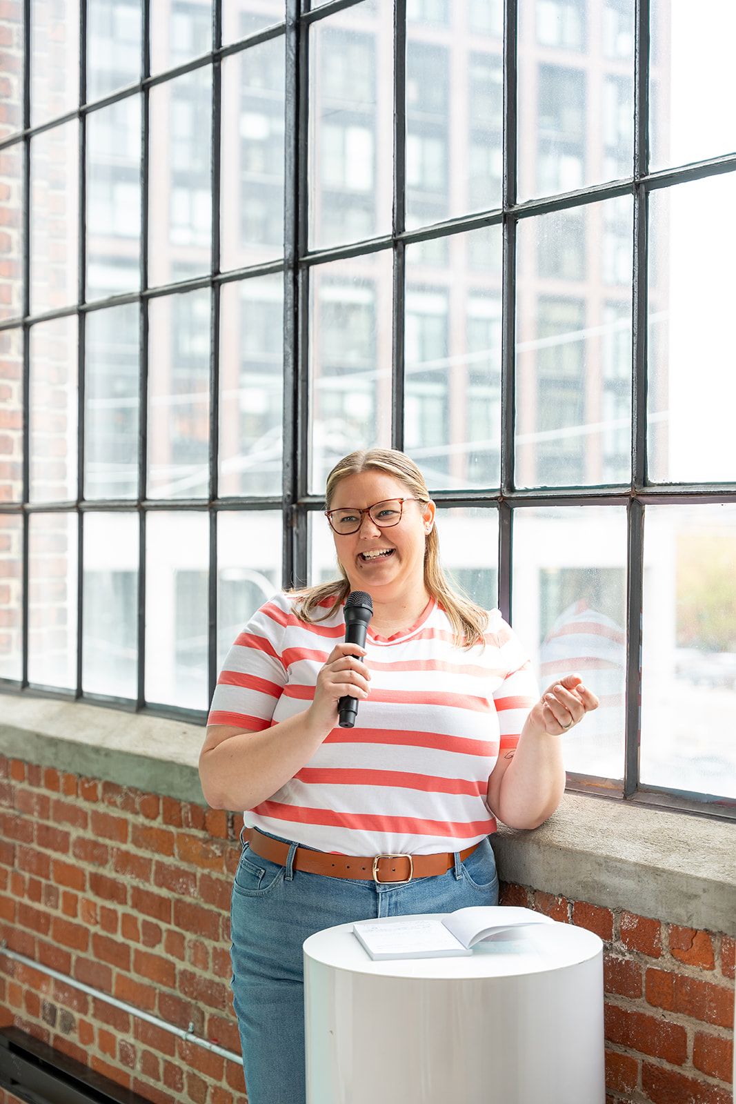 Smiling woman in glasses and a striped top speaks into a microphone beside a white table with an open book near large industrial windows.