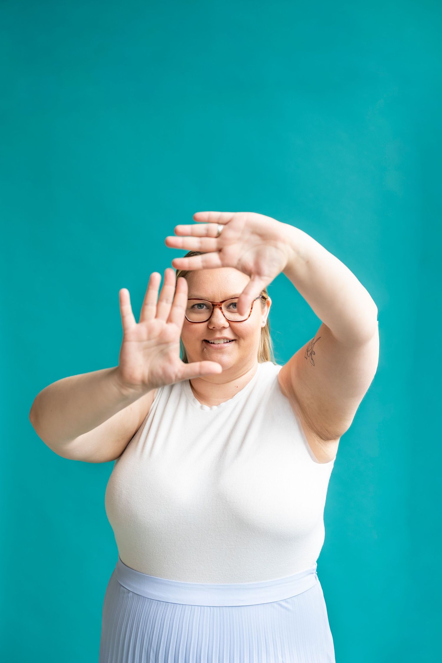 Smiling woman in glasses, white top and pleated skirt frames her face with both hands against a bright teal background.