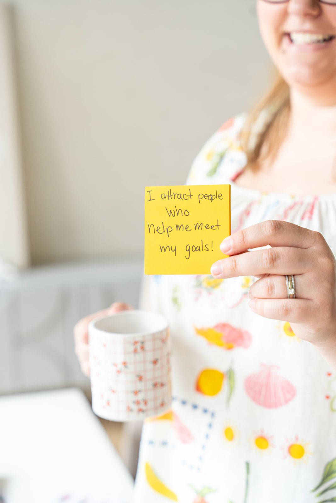 Woman holding yellow sticky note reading 'I attract people who help me meet my goals!' while smiling and holding a coffee mug.