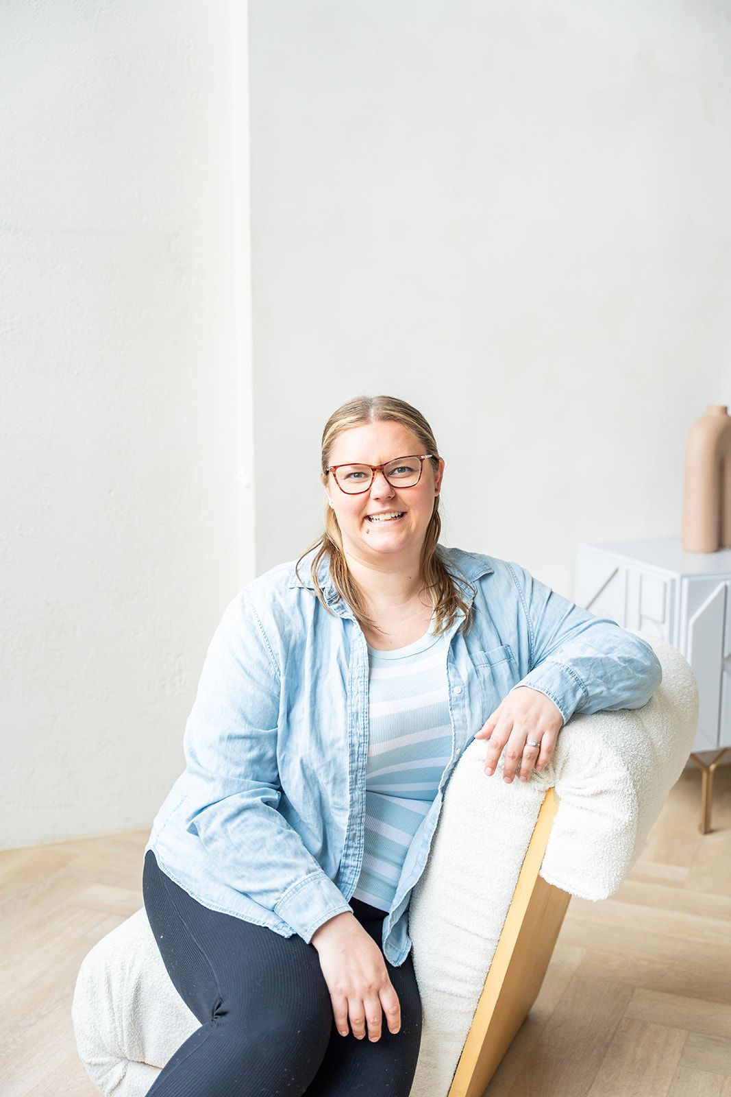 Smiling woman with glasses in light blue shirt sitting relaxed on white armchair in bright minimalist room.