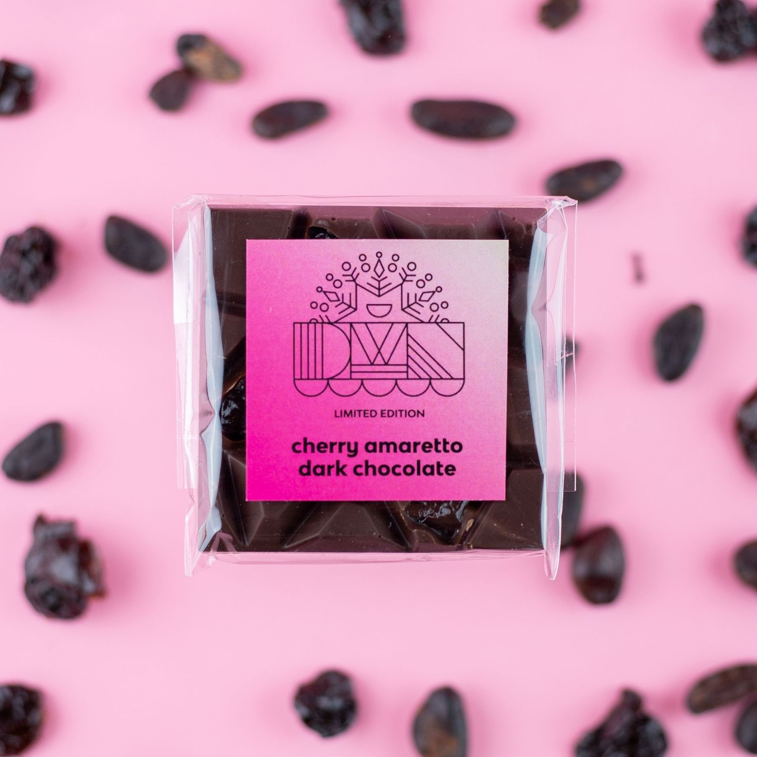 Image of a dark chocolate bar with a cherry amaretto flavor label on a pink background with scattered chocolate pieces.
