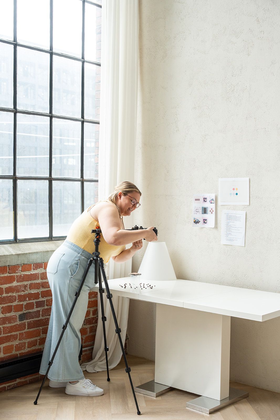 Woman photographer leaning over white table using camera on tripod in bright studio with large industrial windows.