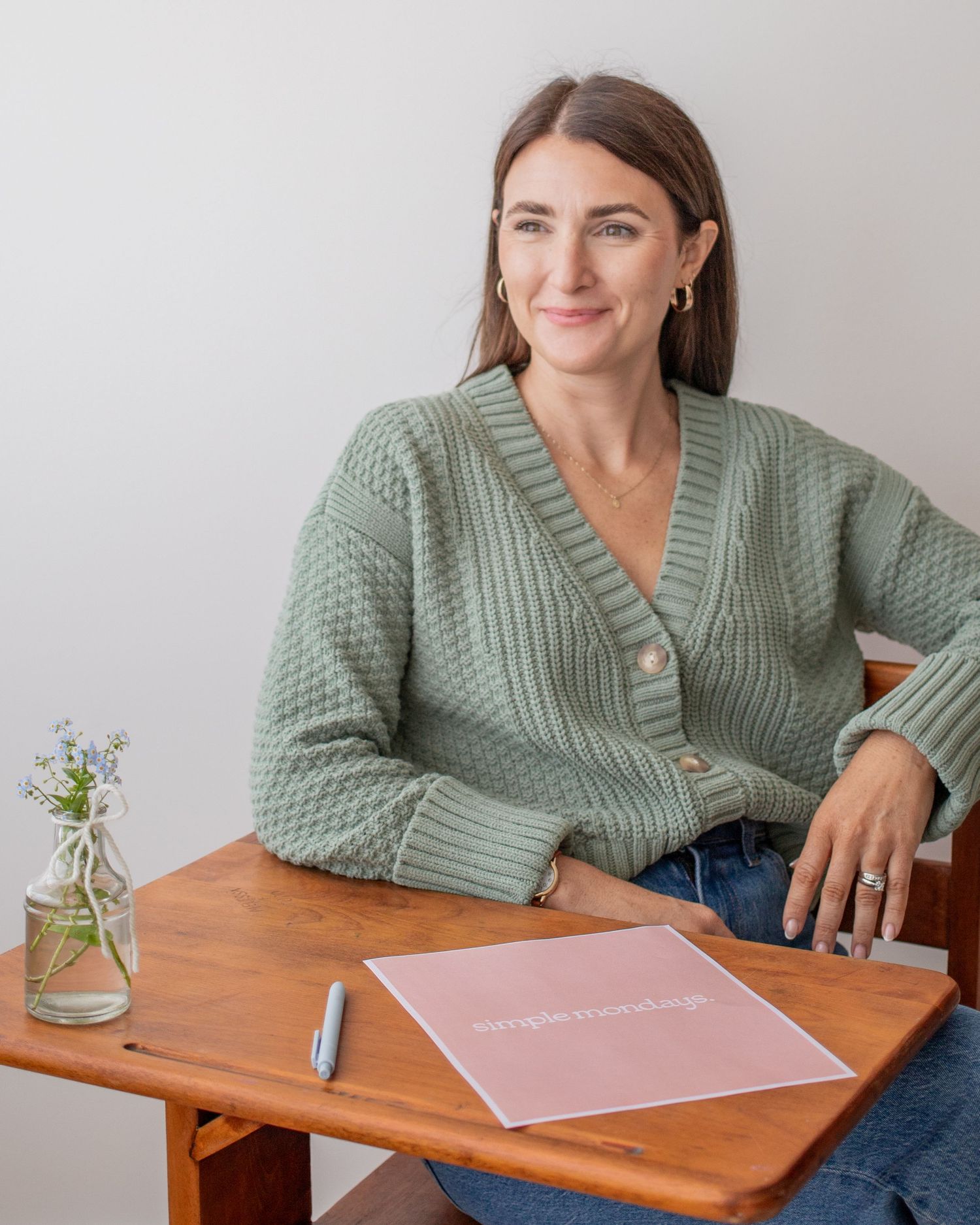 The image shows a smiling woman with long brown hair sitting at a table with various items.