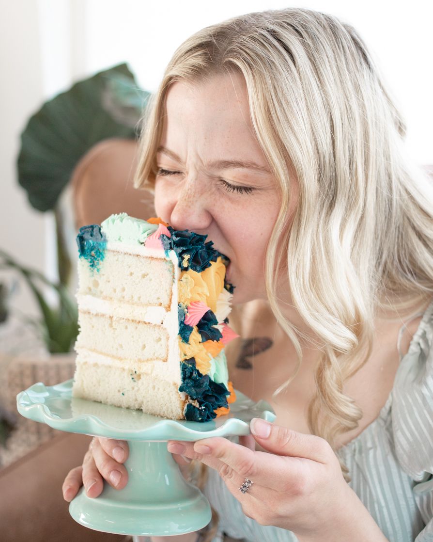 A woman joyfully eats a slice of colorful cake, holding it up to her face.