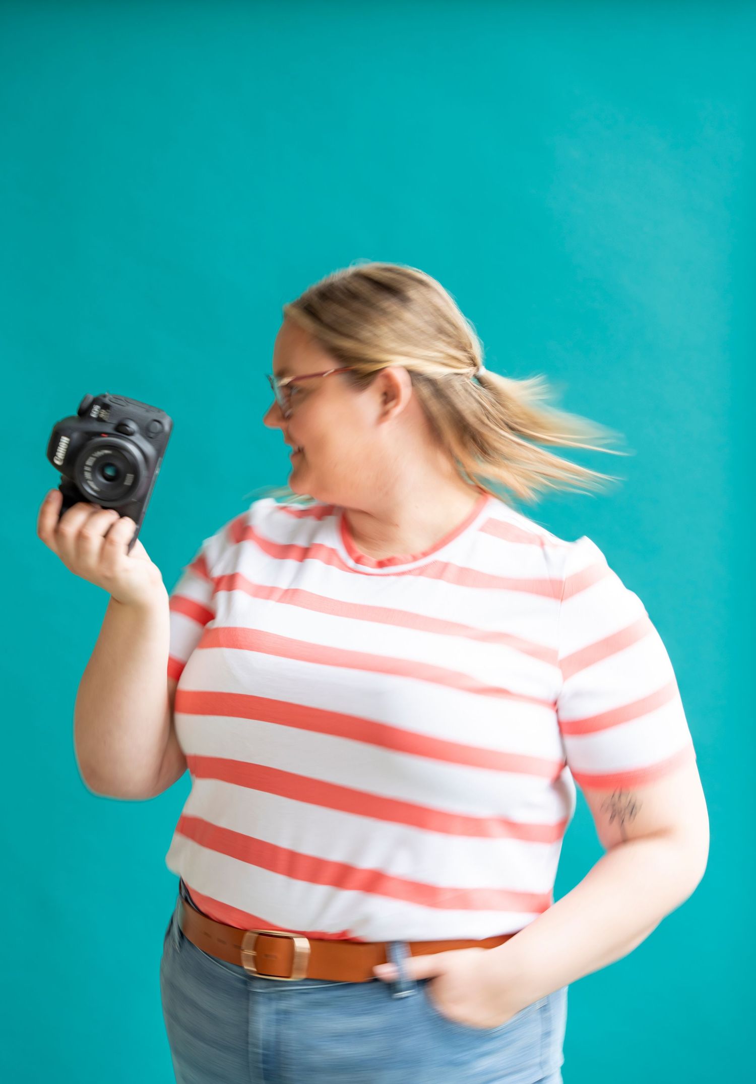 Woman in striped shirt holding a camera against a teal background, smiling and posing for a photo.