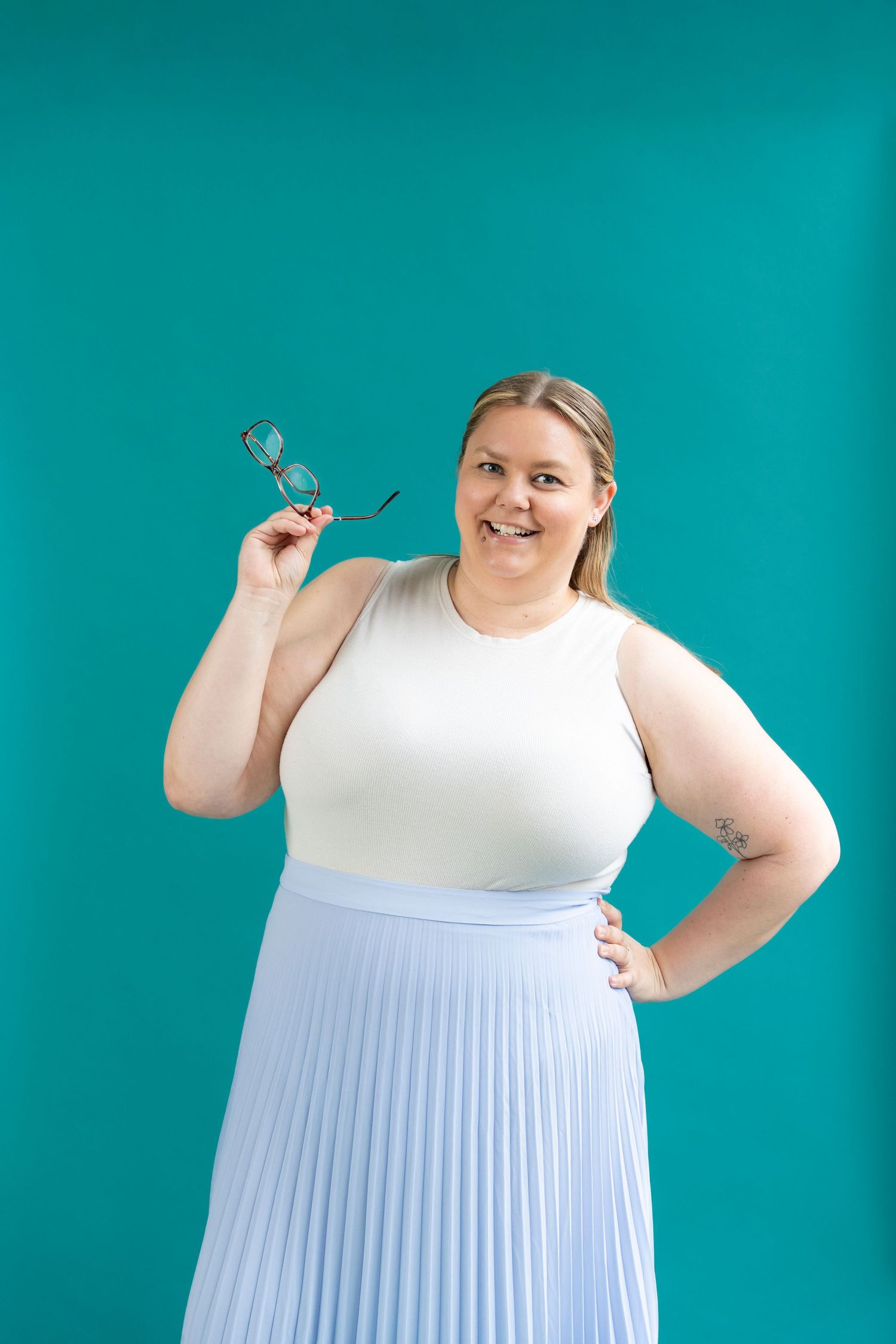 Plus-size woman in white top and blue pleated skirt holding sunglasses, posing on teal background.