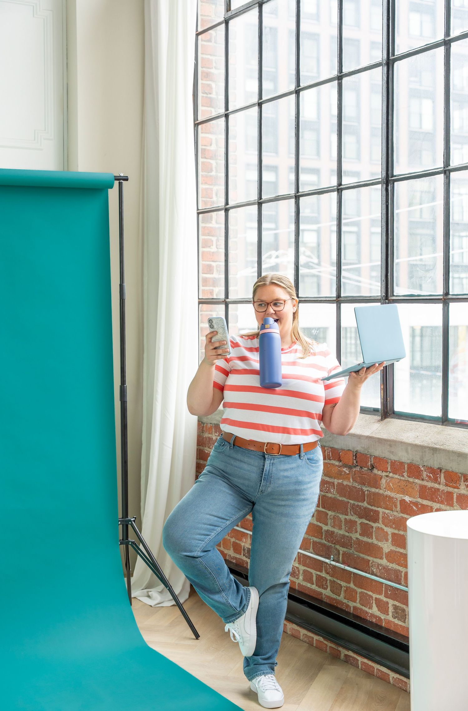 Woman in striped shirt holding water bottle, phone, and laptop near large industrial windows with teal backdrop.