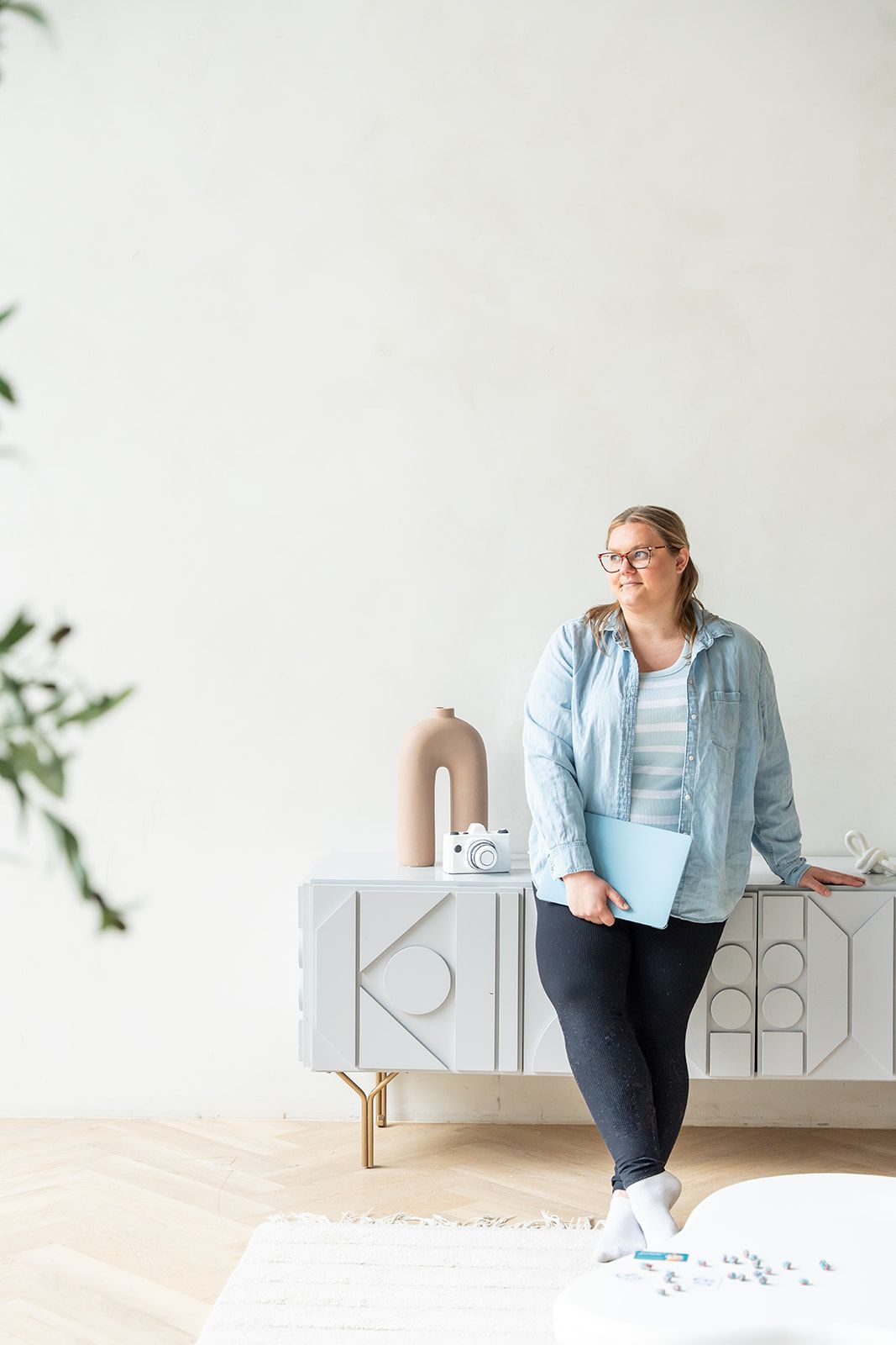 Woman in glasses holding blue folder stands by modern white sideboard in bright minimalist living room.