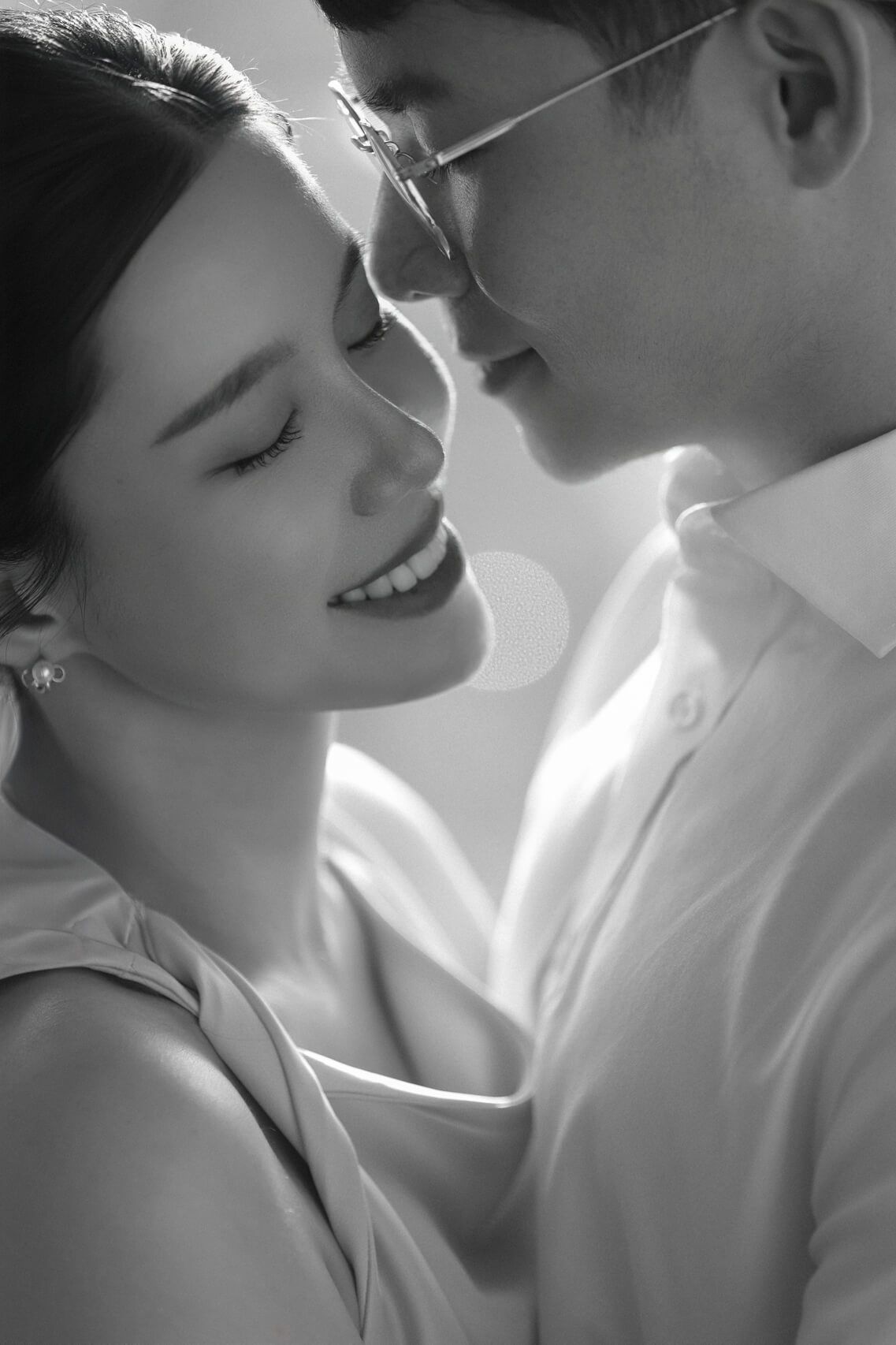 An intimate black and white wedding photograph capturing an Asian couple's quiet strength and connection. Shot in natural, backlit lighting