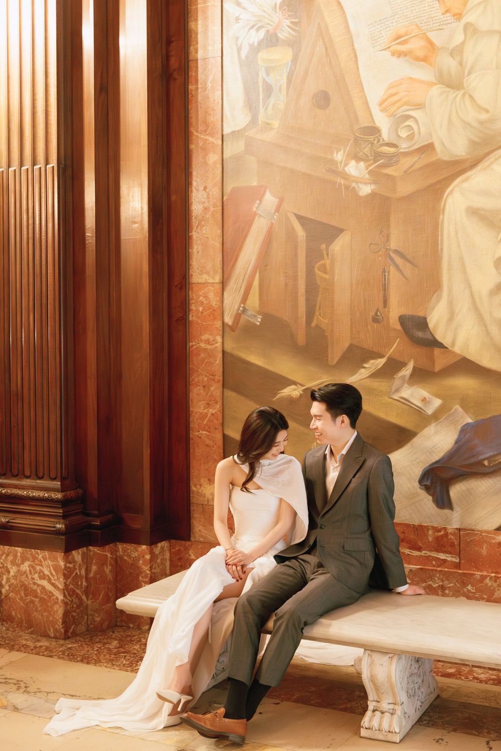 An asian couple sits on a marble bench sharing smiles in front of a grand mural during their timeless New York engagement session at NYPL.