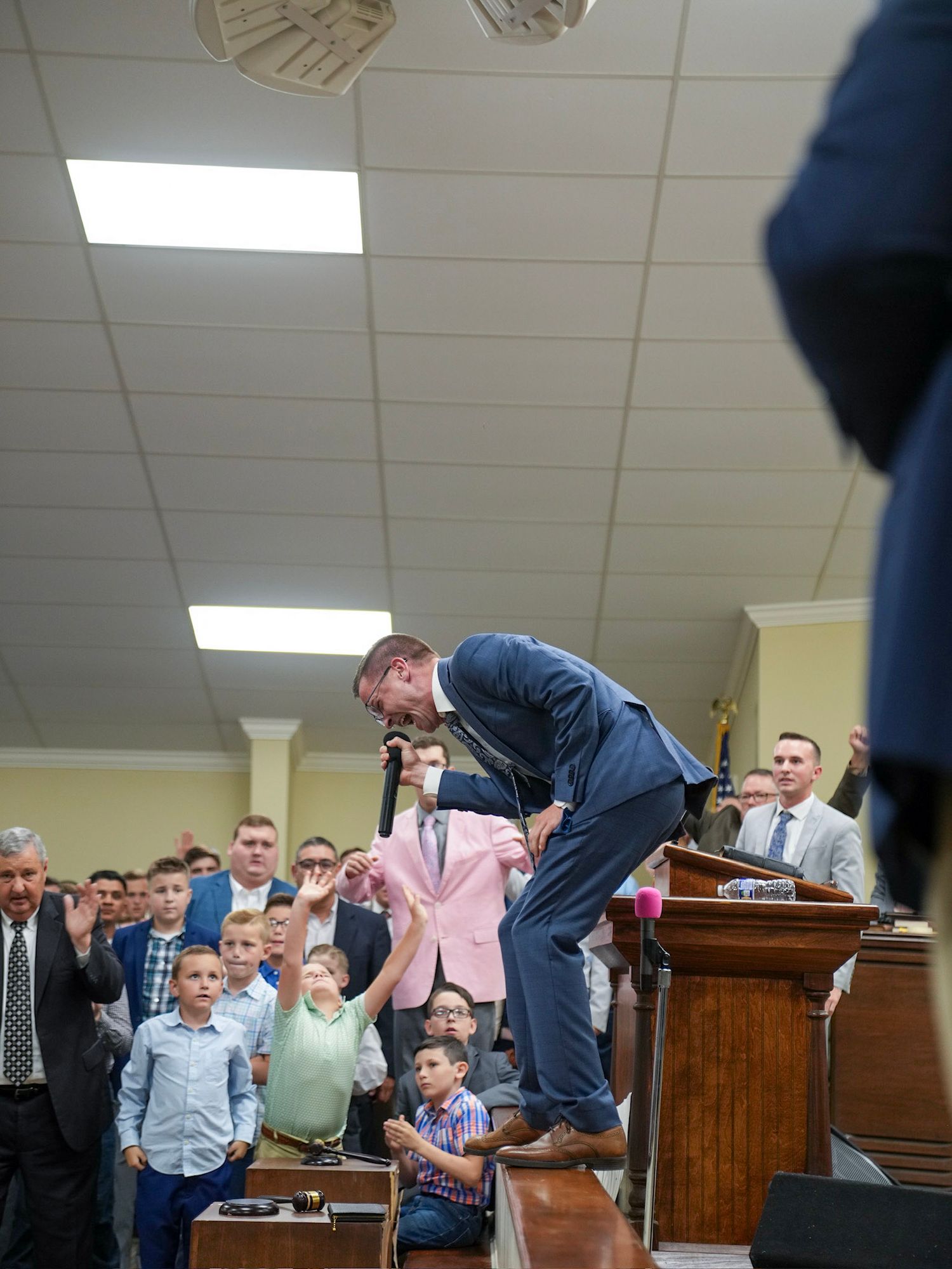 A preacher holds a pink blazer while standing at a pulpit before a congregation of children and adults in a church.
