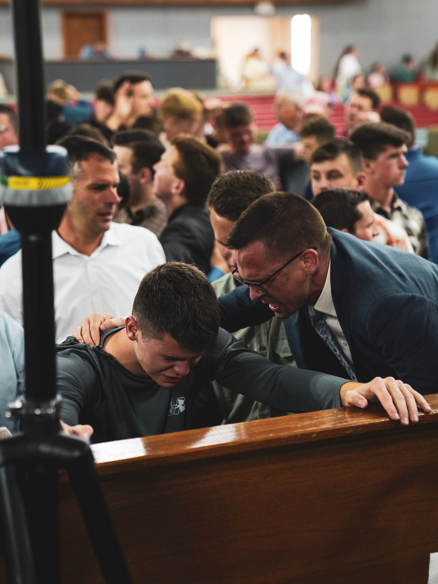 A man in a suit comforts a crying young man at a church pew during a religious service.