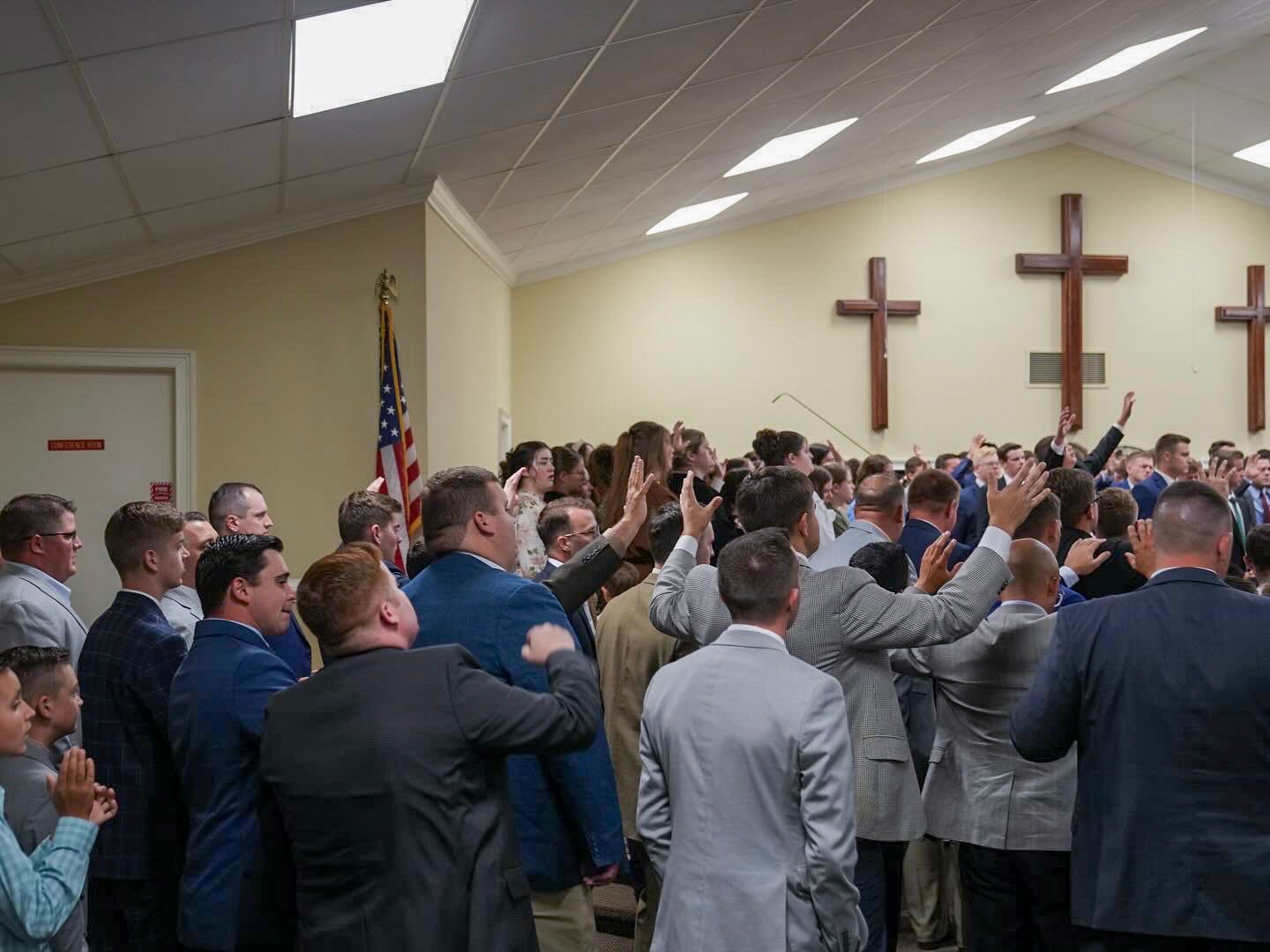 Men in suits raising hands in prayer before two wooden crosses in church.