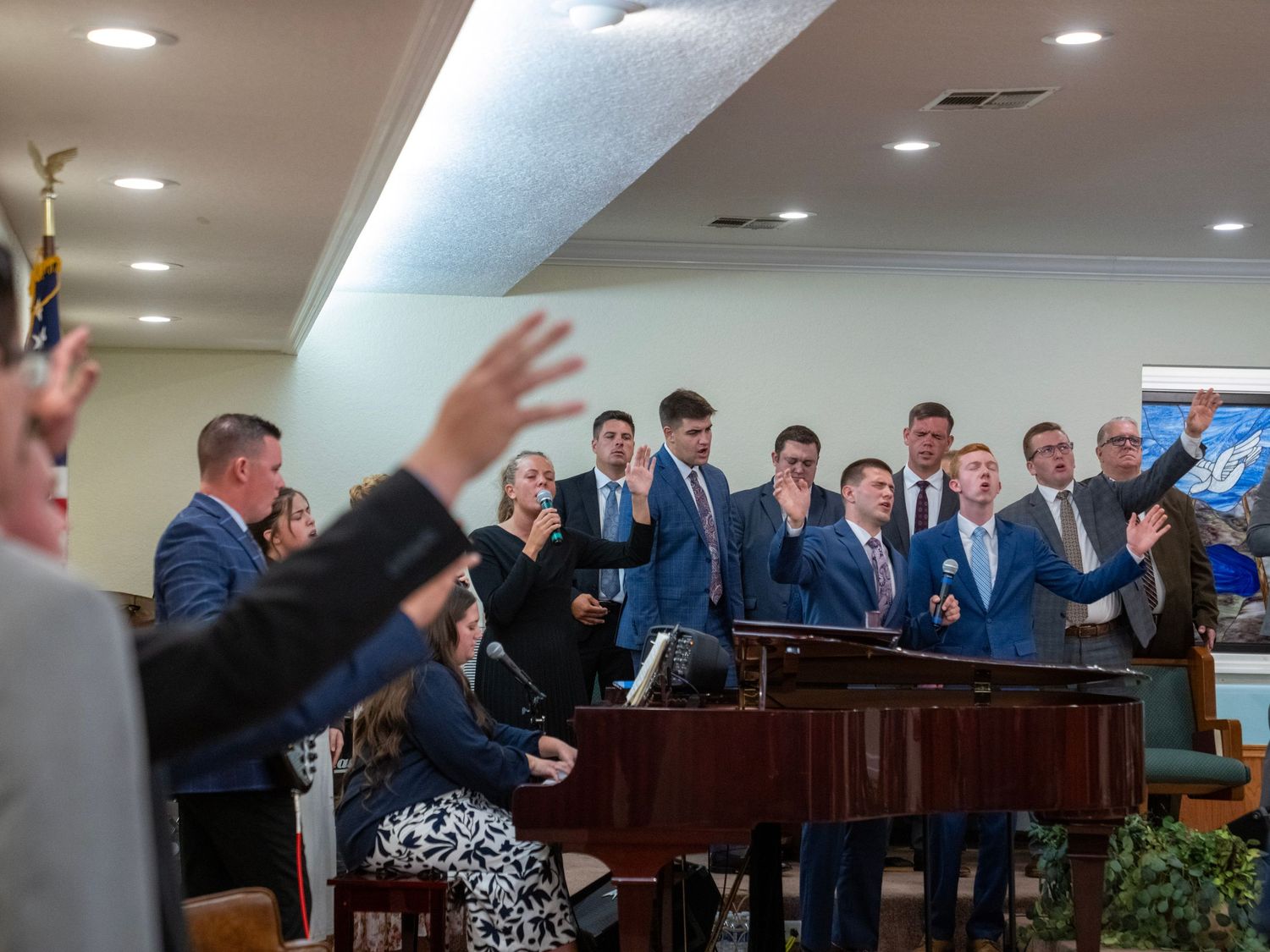 Choir group in blue suits singing around a piano in a church worship service.