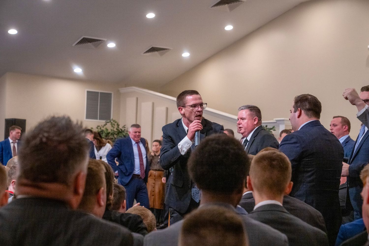 Man speaking into microphone at church event surrounded by men in suits in decorated venue.