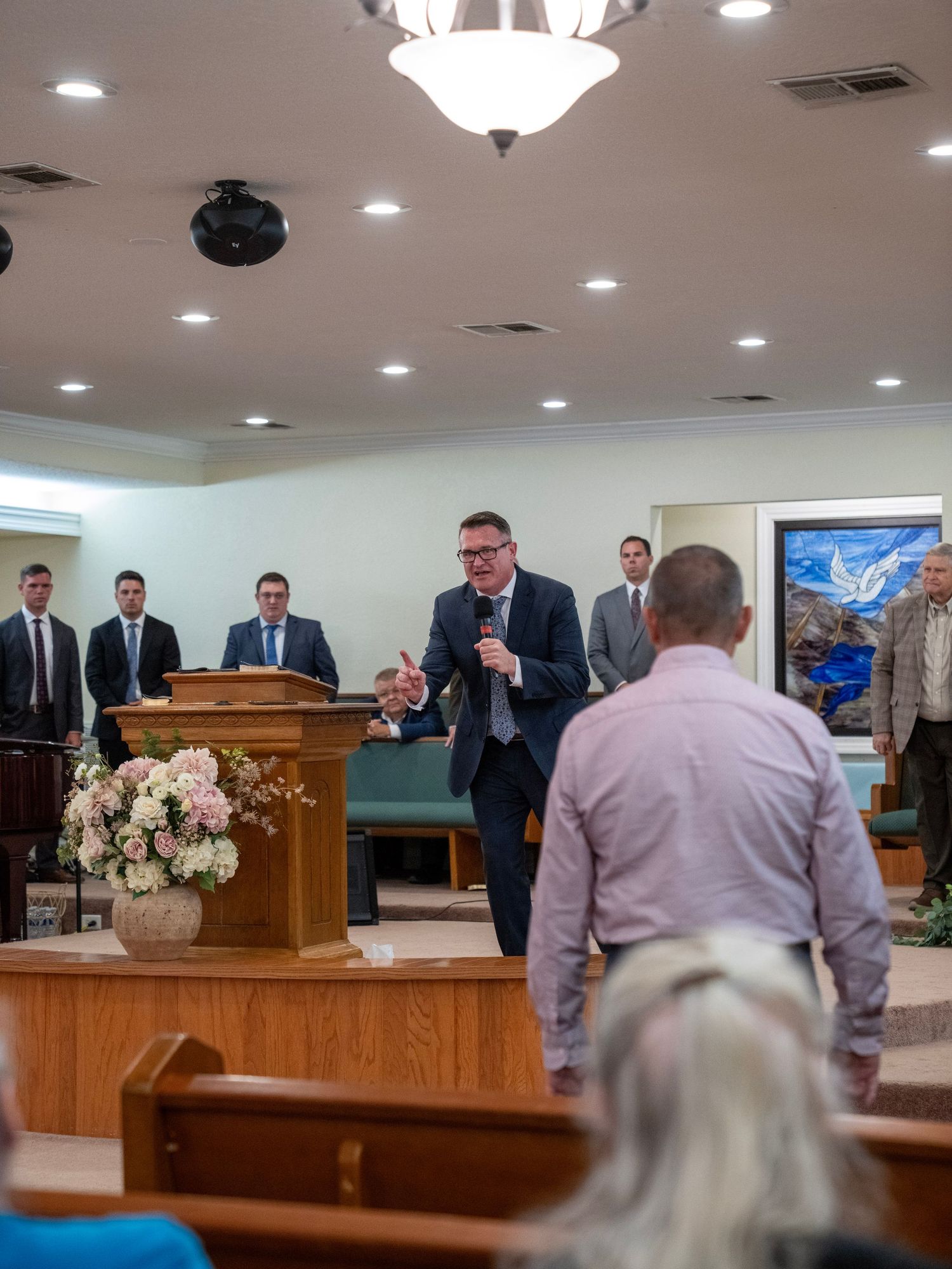 Pastor speaking at a church pulpit with congregation members standing nearby.