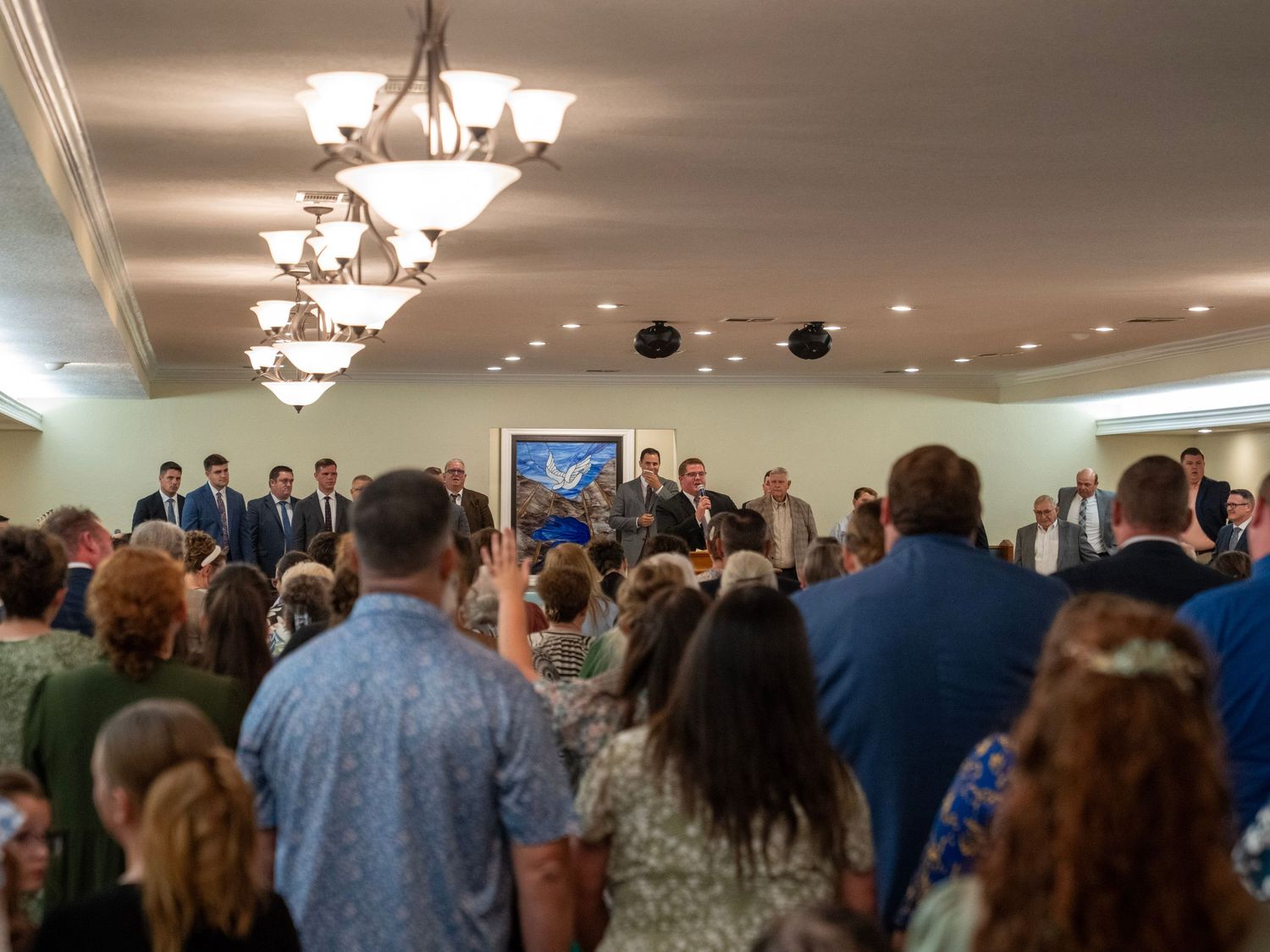 Large congregation standing in worship with hands raised in an elegant chandelier-lit church hall.