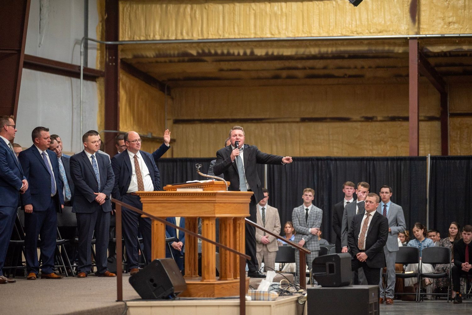 A preacher delivers a passionate sermon at a pulpit surrounded by men in suits inside a large warehouse church.
