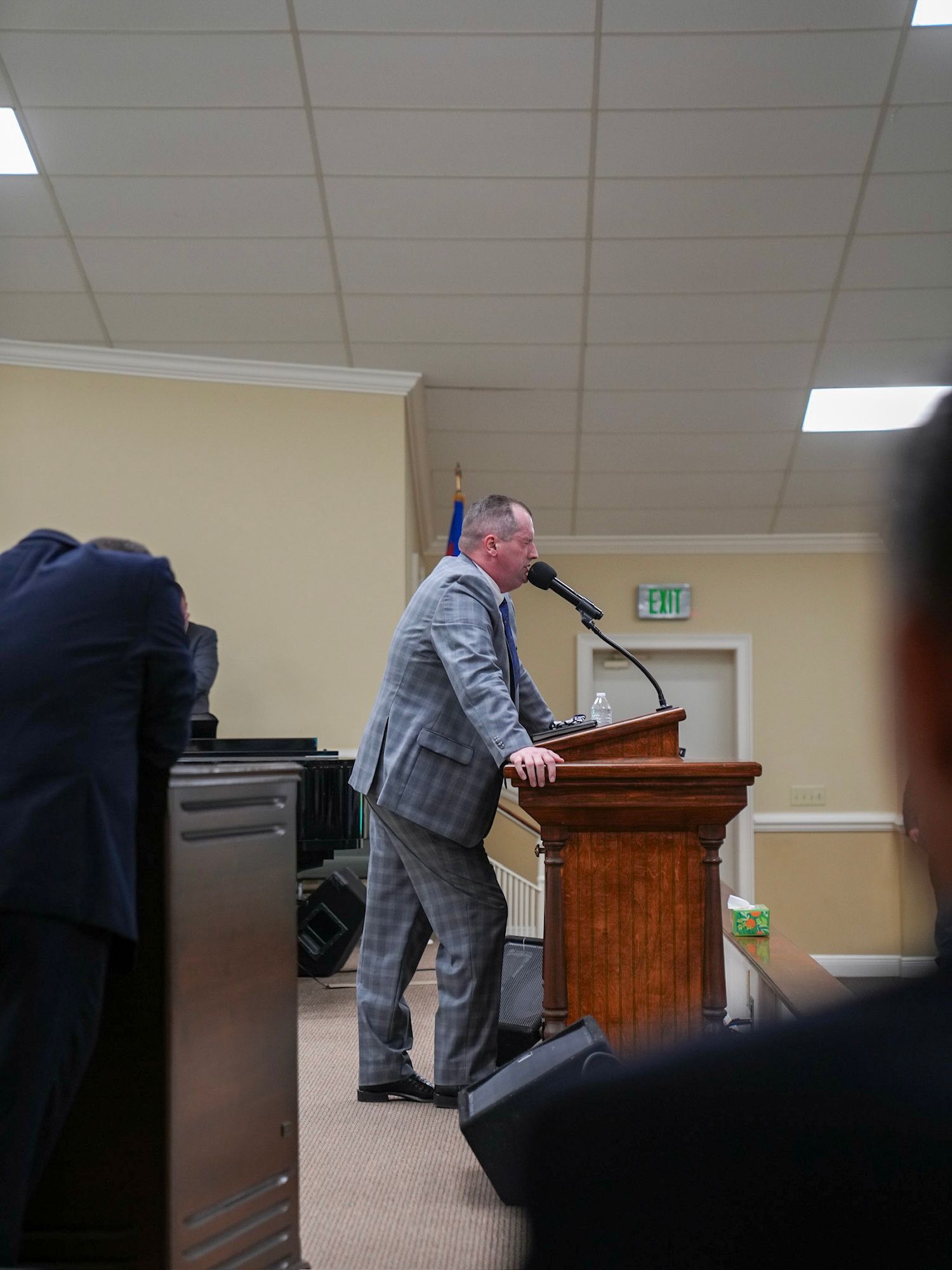 Man in plaid suit delivering speech at wooden podium with microphone in church setting.