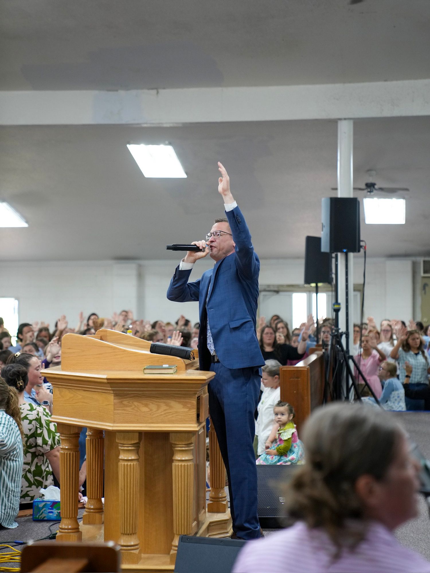 A pastor in a blue suit raises his hand while preaching with a microphone at a church pulpit.