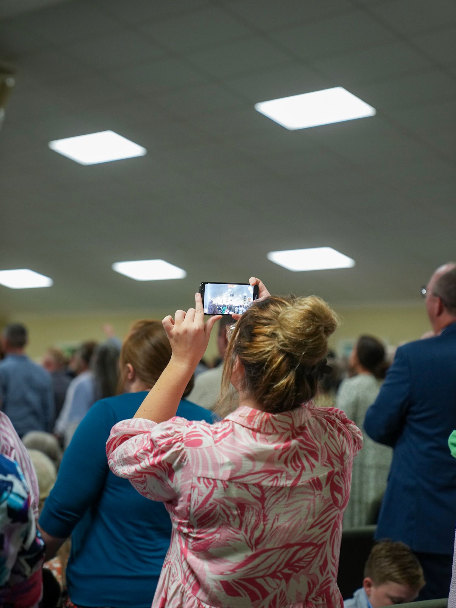 Woman in floral dress photographing church congregation during religious service.